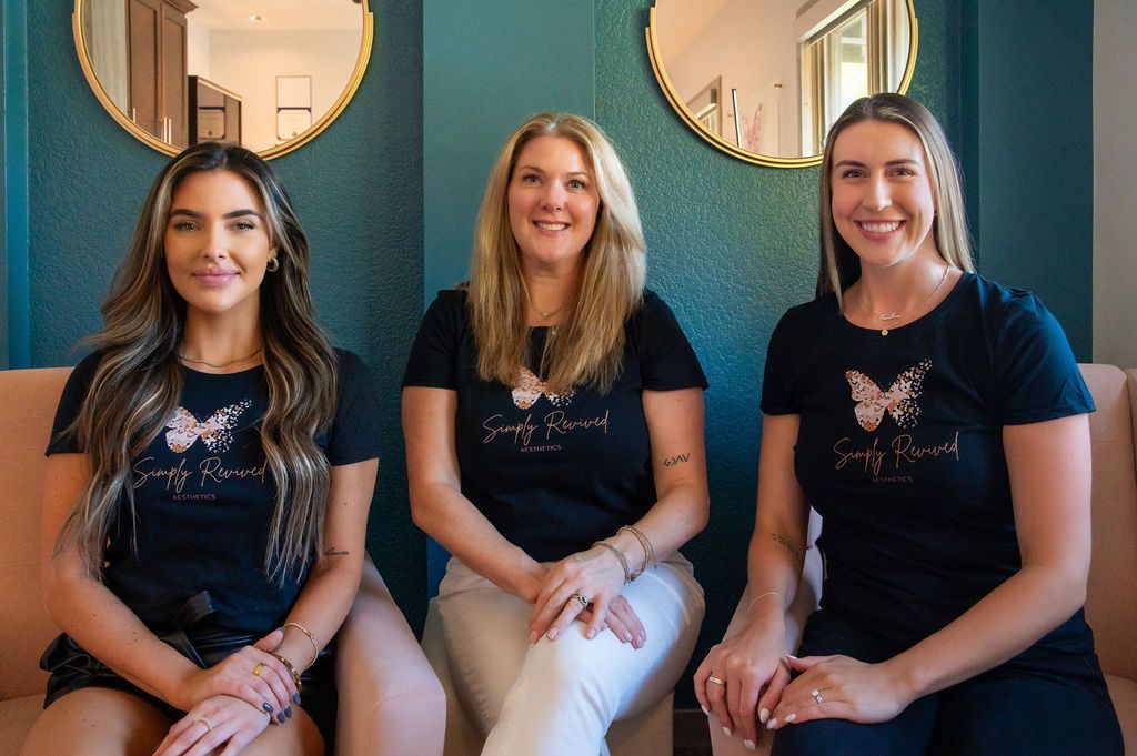Three women in black shirts with a butterfly logo, sitting on a pink couch. Blue wall background.