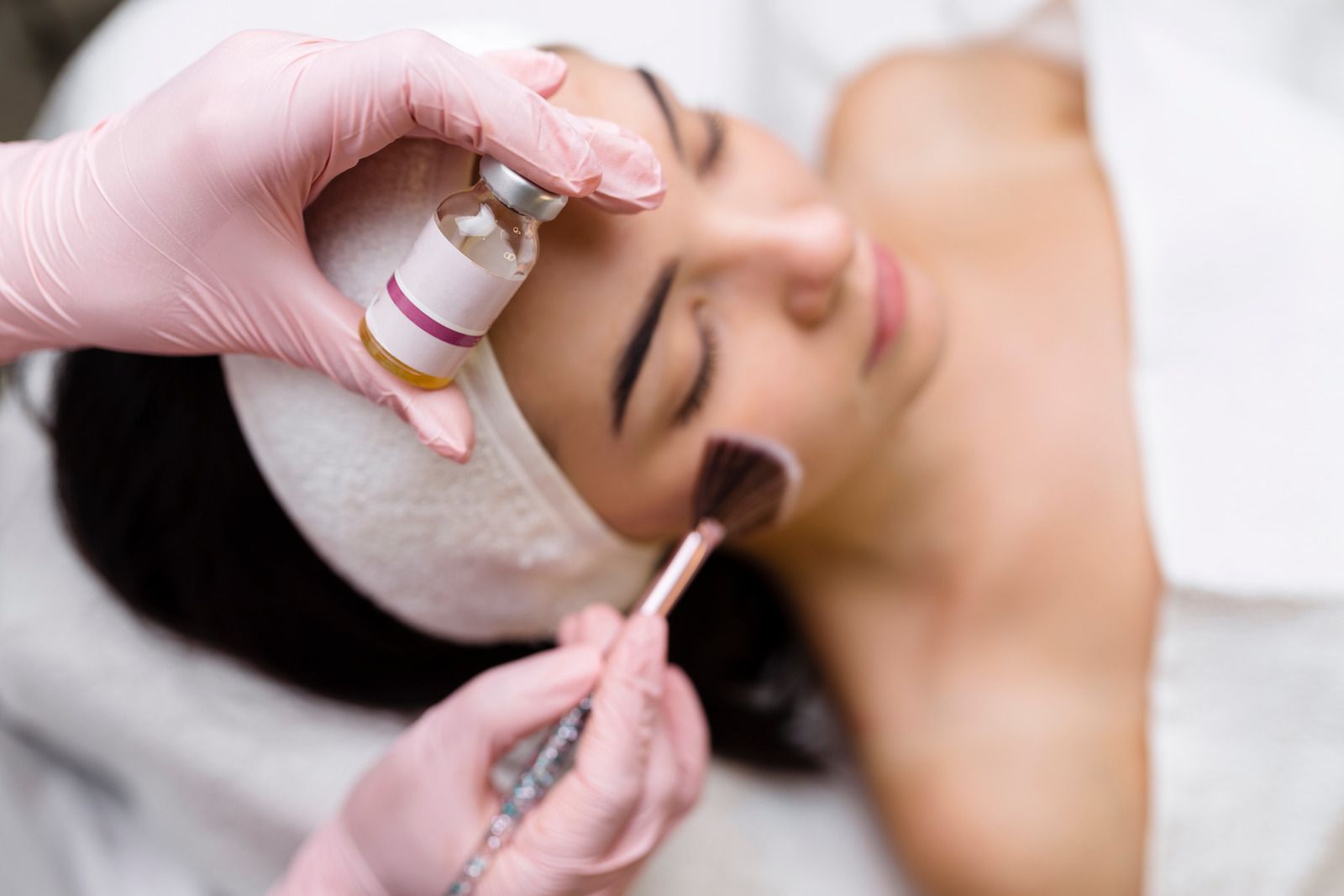 Gloved hands apply a facial treatment and makeup brush to a woman lying on a spa bed
