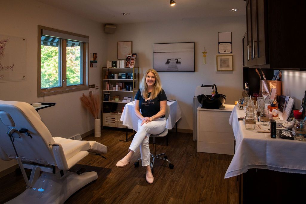 Woman sitting in a bright room; desk and treatment chair. She wears a dark shirt and white pants, smiling.