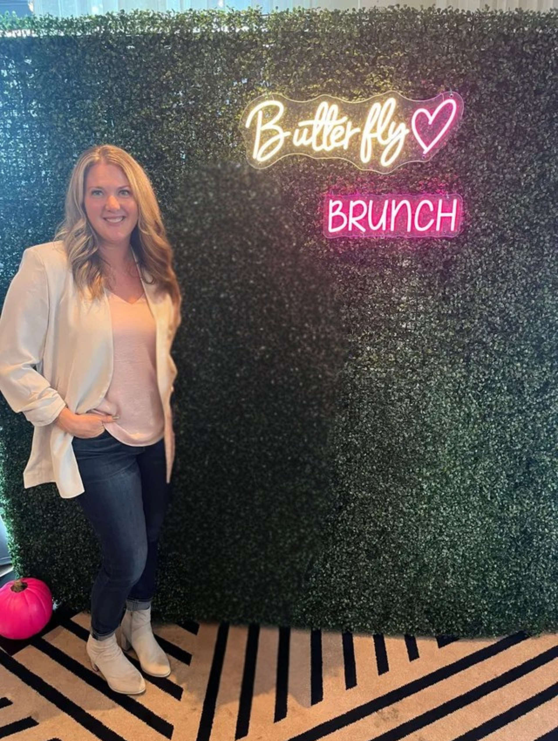 Woman at Butterfly Brunch with neon sign on a green wall, hands in pockets, wearing white blazer and jeans.