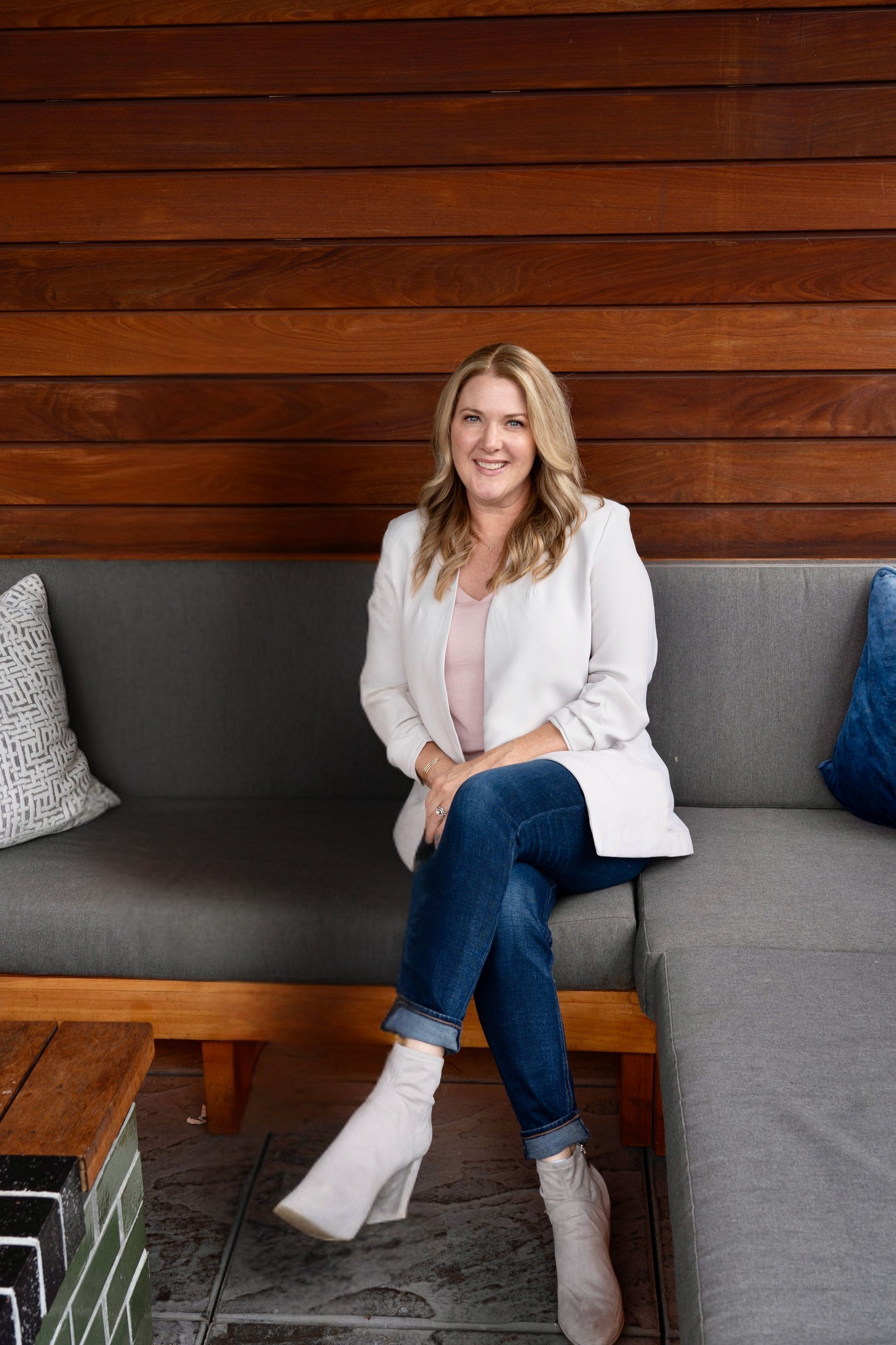 Woman seated on a gray outdoor sofa, wearing a white blazer, jeans, and white boots, smiling.