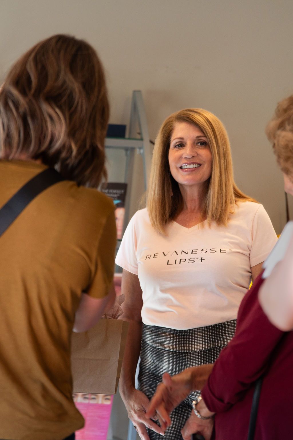Woman in white t-shirt smiles at two others. One is facing her, the other is on the right. Indoors.