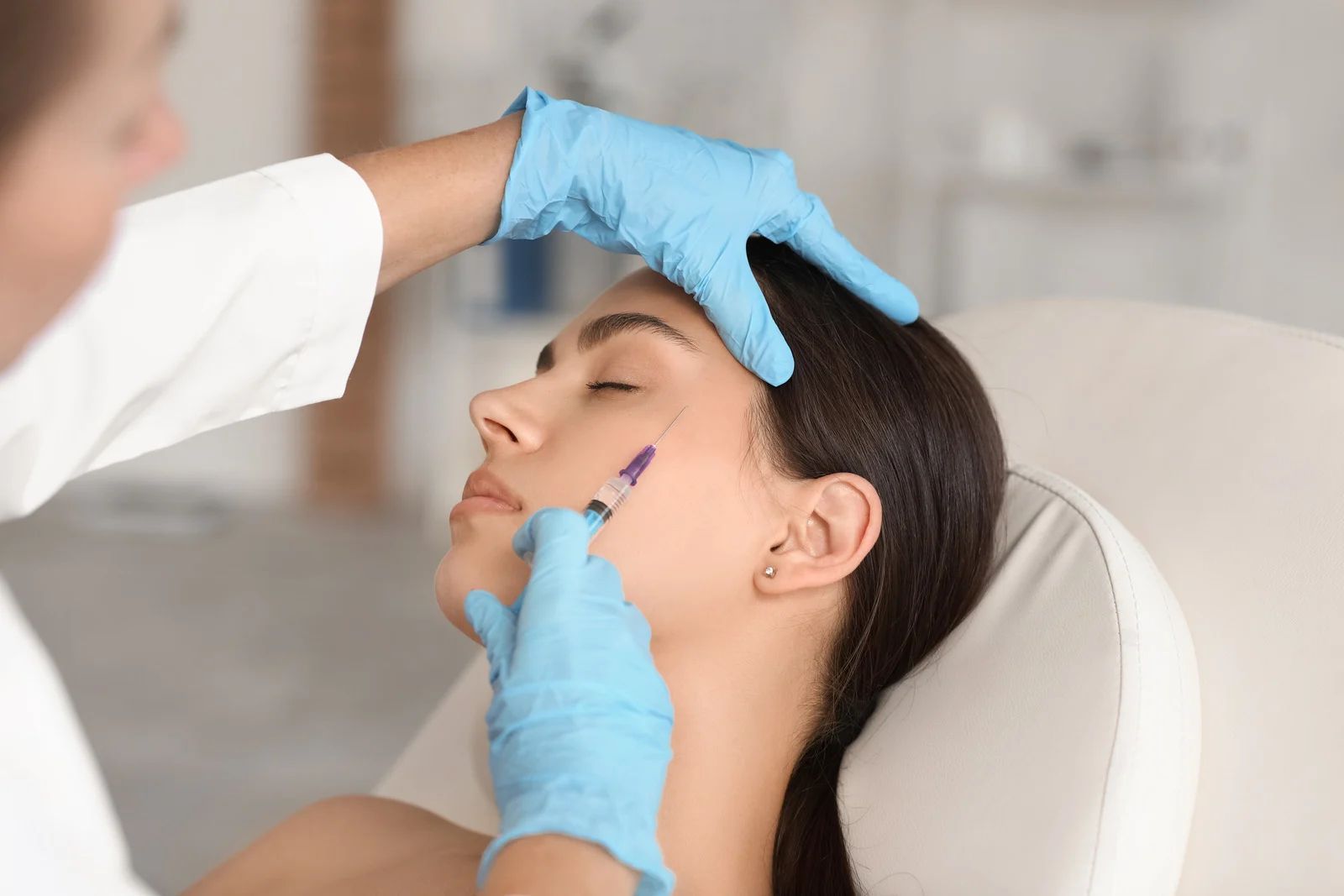 A medical professional in gloves administers a facial filler injection to a person's temple area in a clinic.