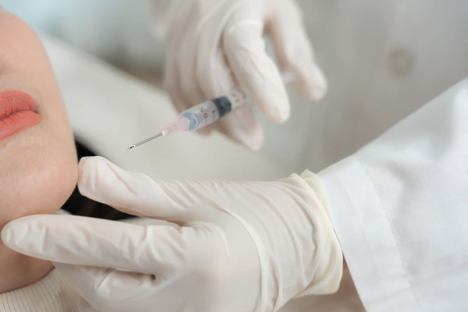 A gloved hand injecting a syringe into a person's jawline, in a medical setting.
