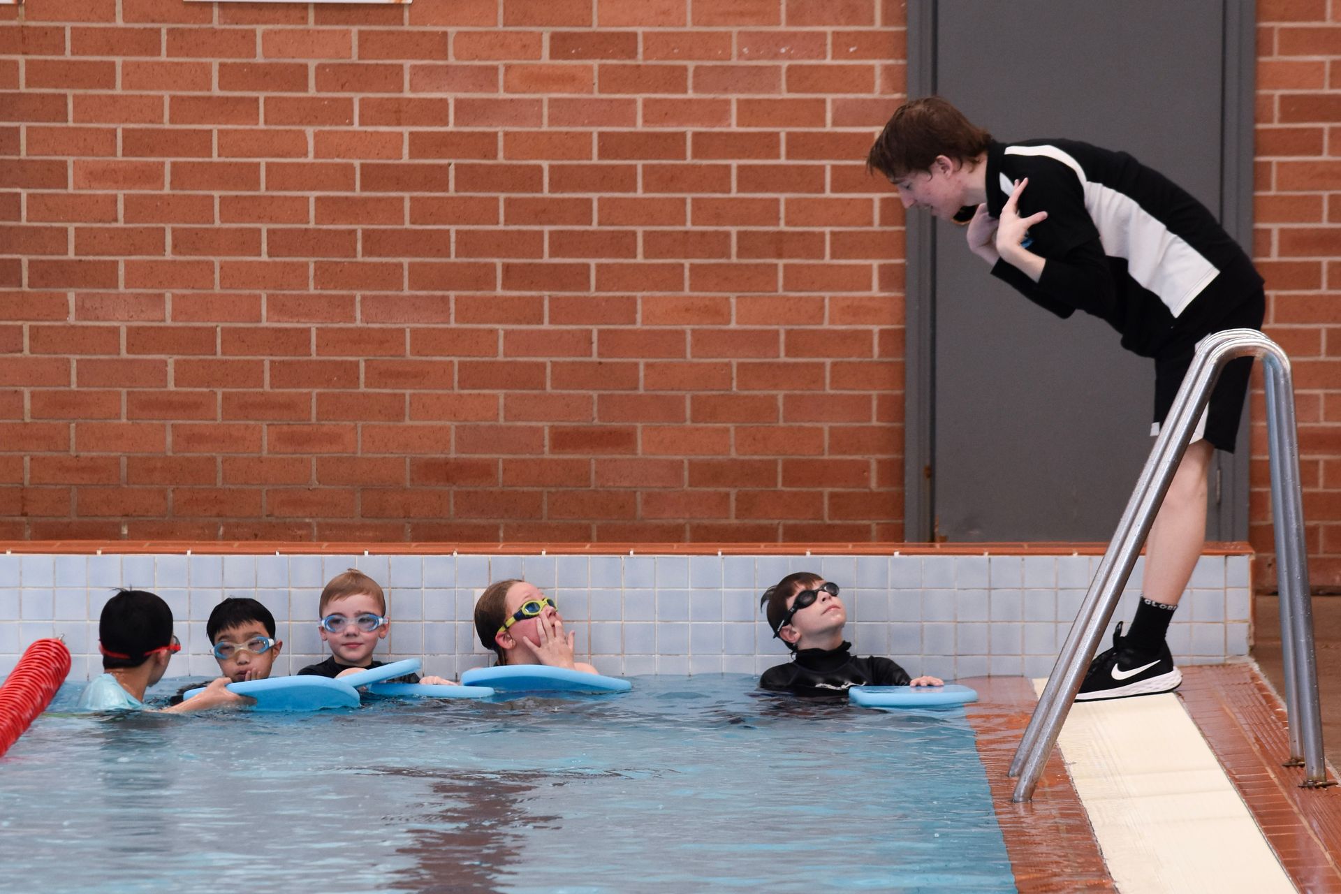 A group of children are swimming in a pool while a man stands on a ladder.