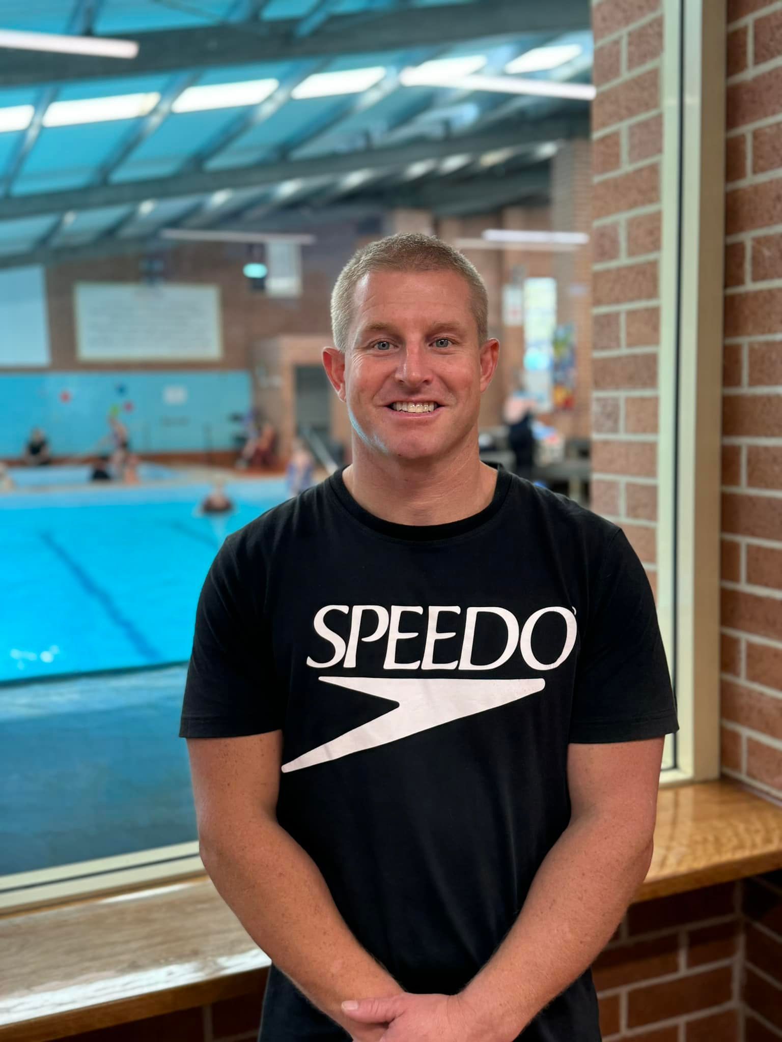 A man wearing a speedo shirt is standing in front of a swimming pool.