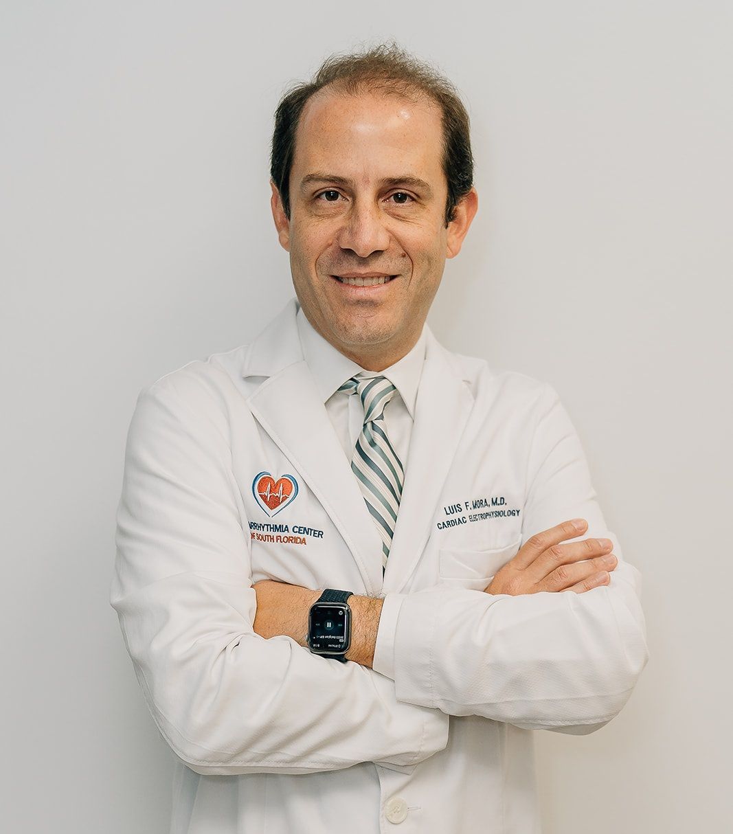 Doctor in a white coat with arms crossed, standing against a light gray background.