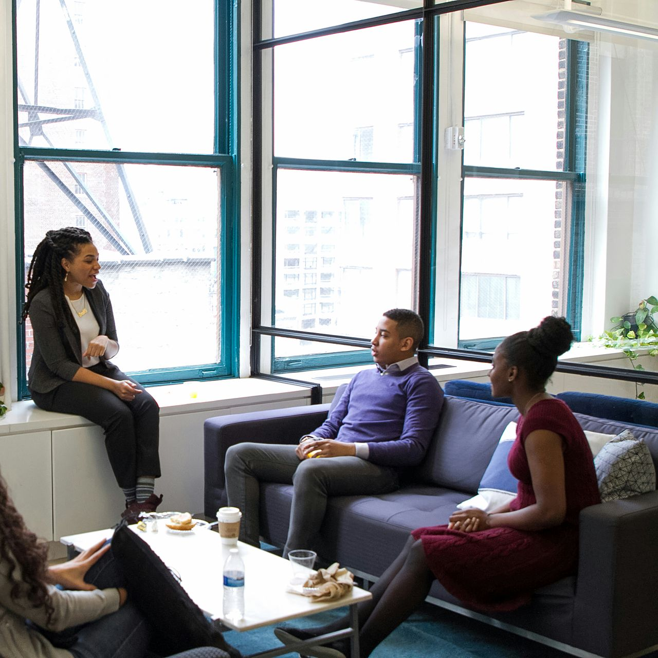 Group of people in an office lounge, some seated on a couch, others on a windowsill, engaged in conversation.