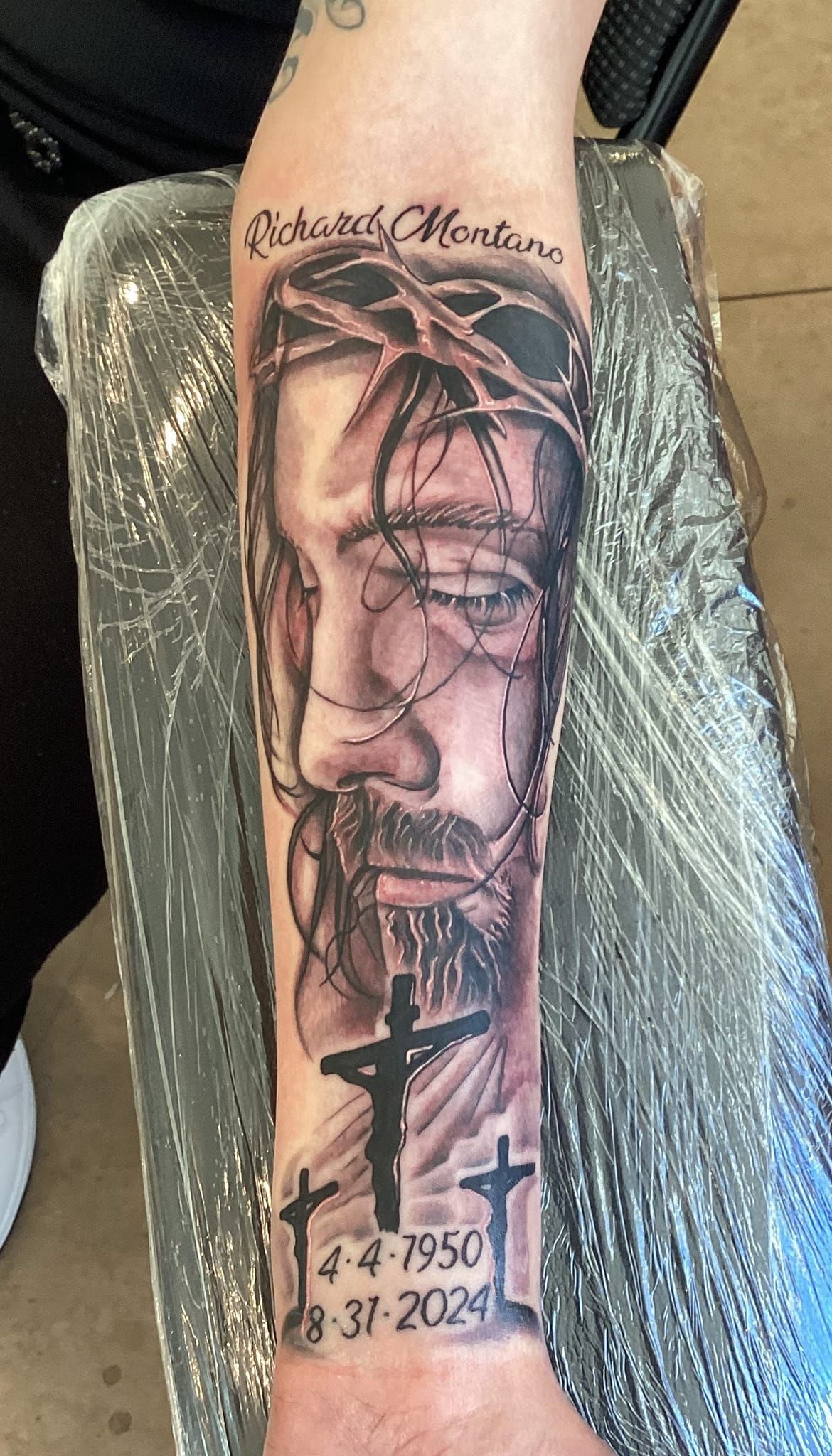 Black and white tattoo of Jesus wearing a crown of thorns on an arm, with three crosses and dates.