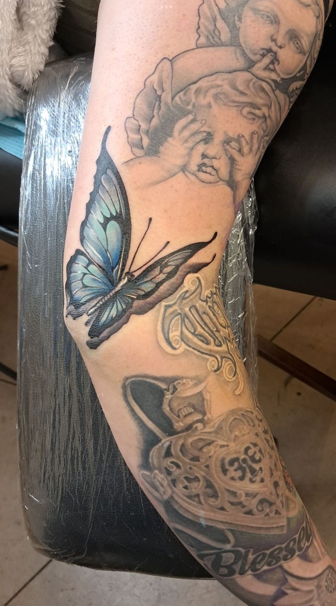 Tattoo of a blue butterfly with pixelated wings on an arm, with other detailed grayscale tattoos.