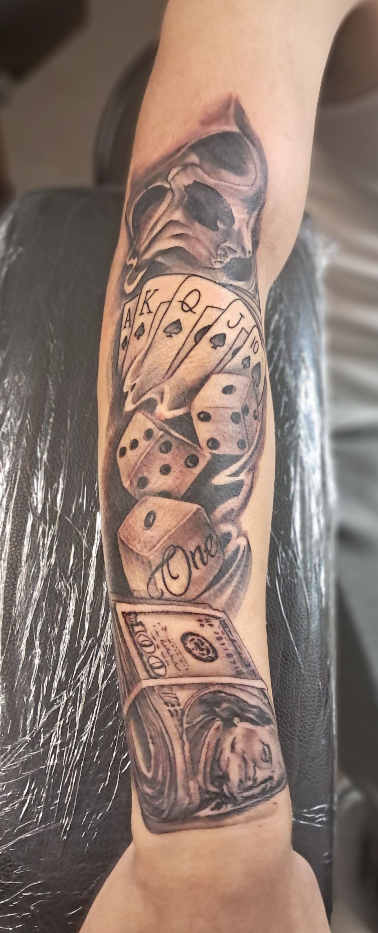 Black and gray forearm tattoo of dice, a skull, and a dollar bill.