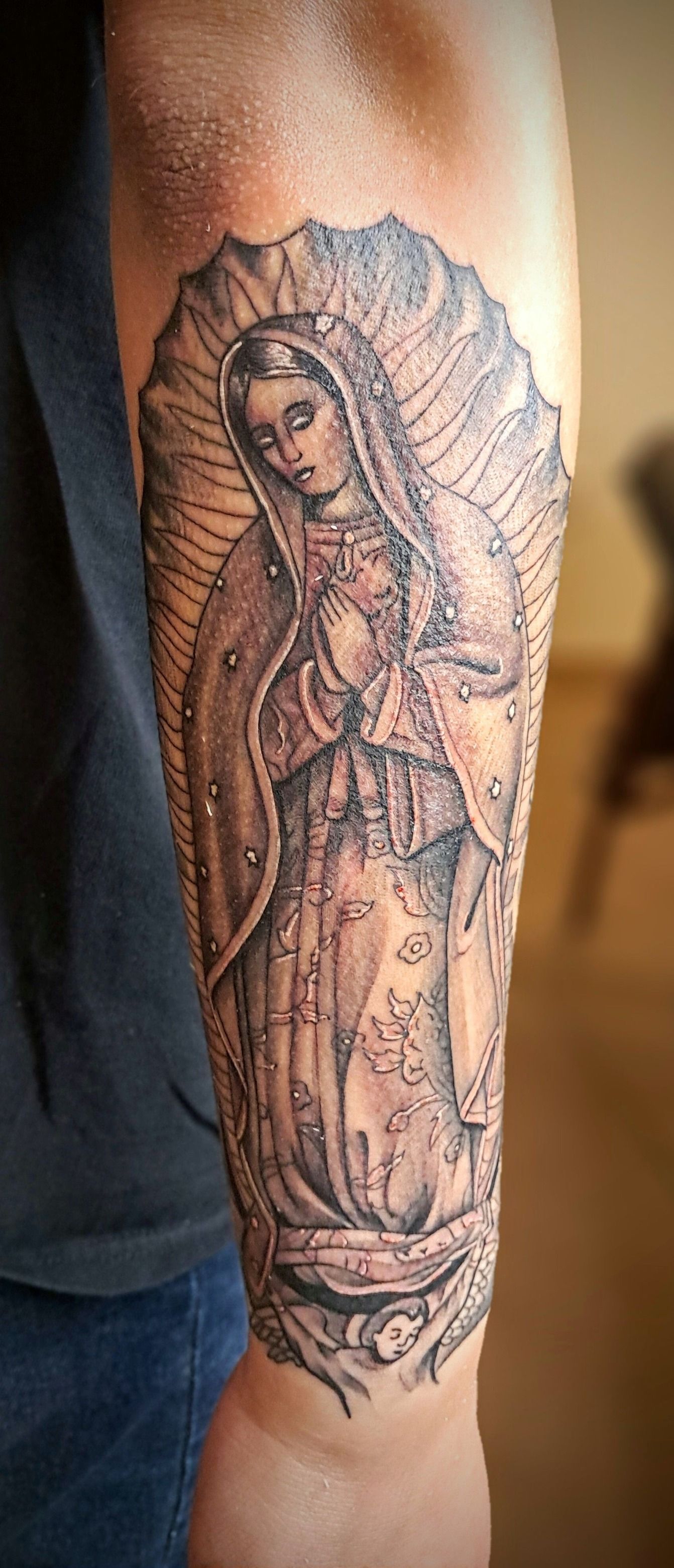 Tattoo of the Virgin Mary on an arm, in black and gray tones.