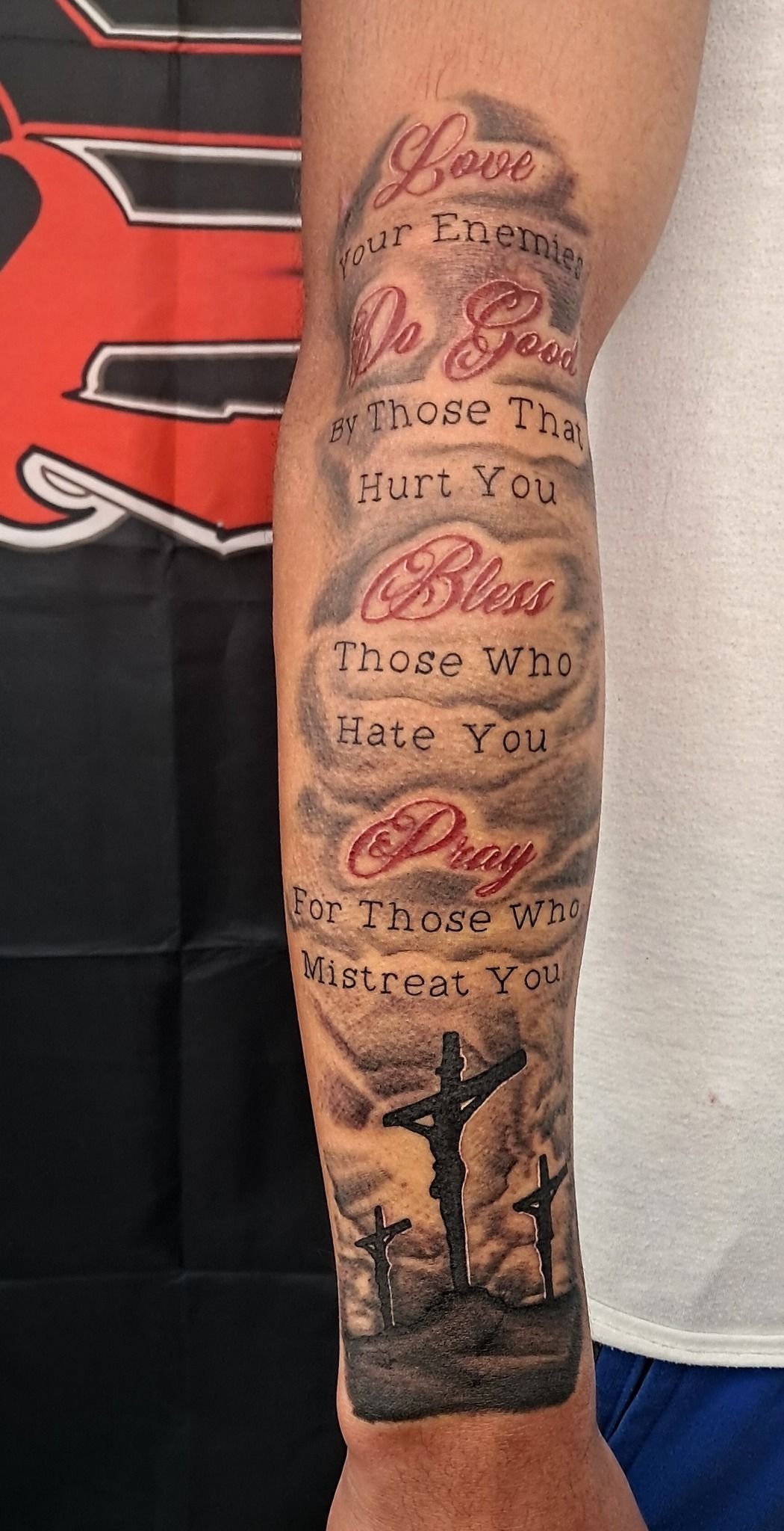 Tattoo of a biblical verse on a forearm. Text in clouds above three crosses under a stormy sky.