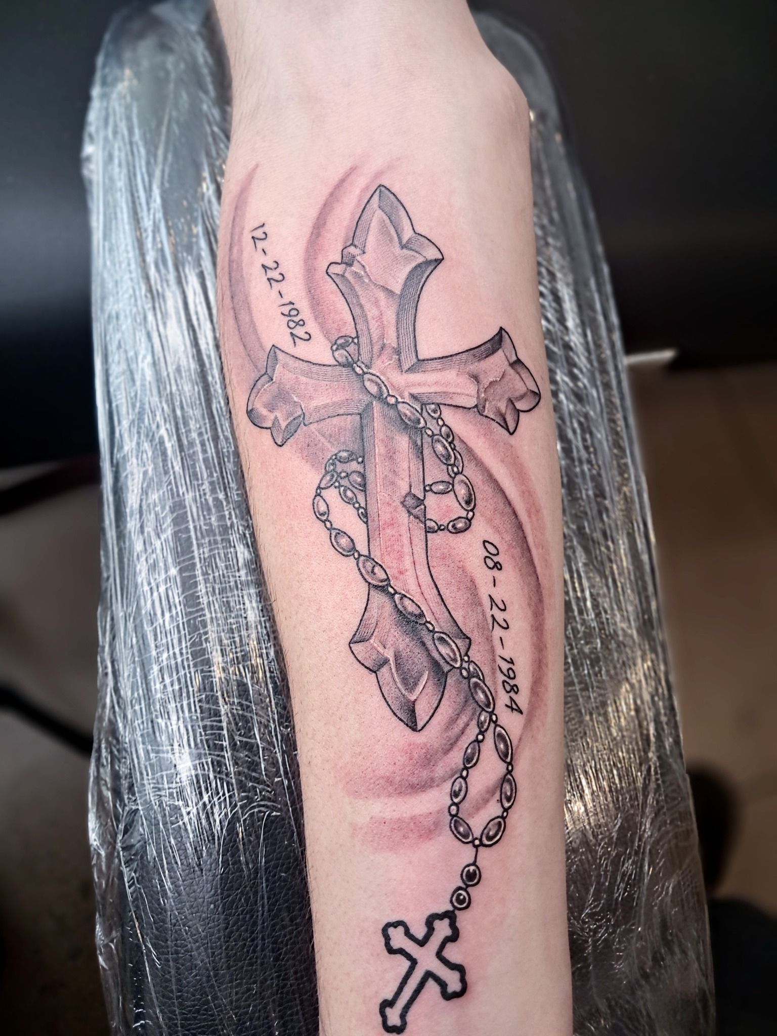 Cross tattoo on arm, black ink, with rosary beads and dates.