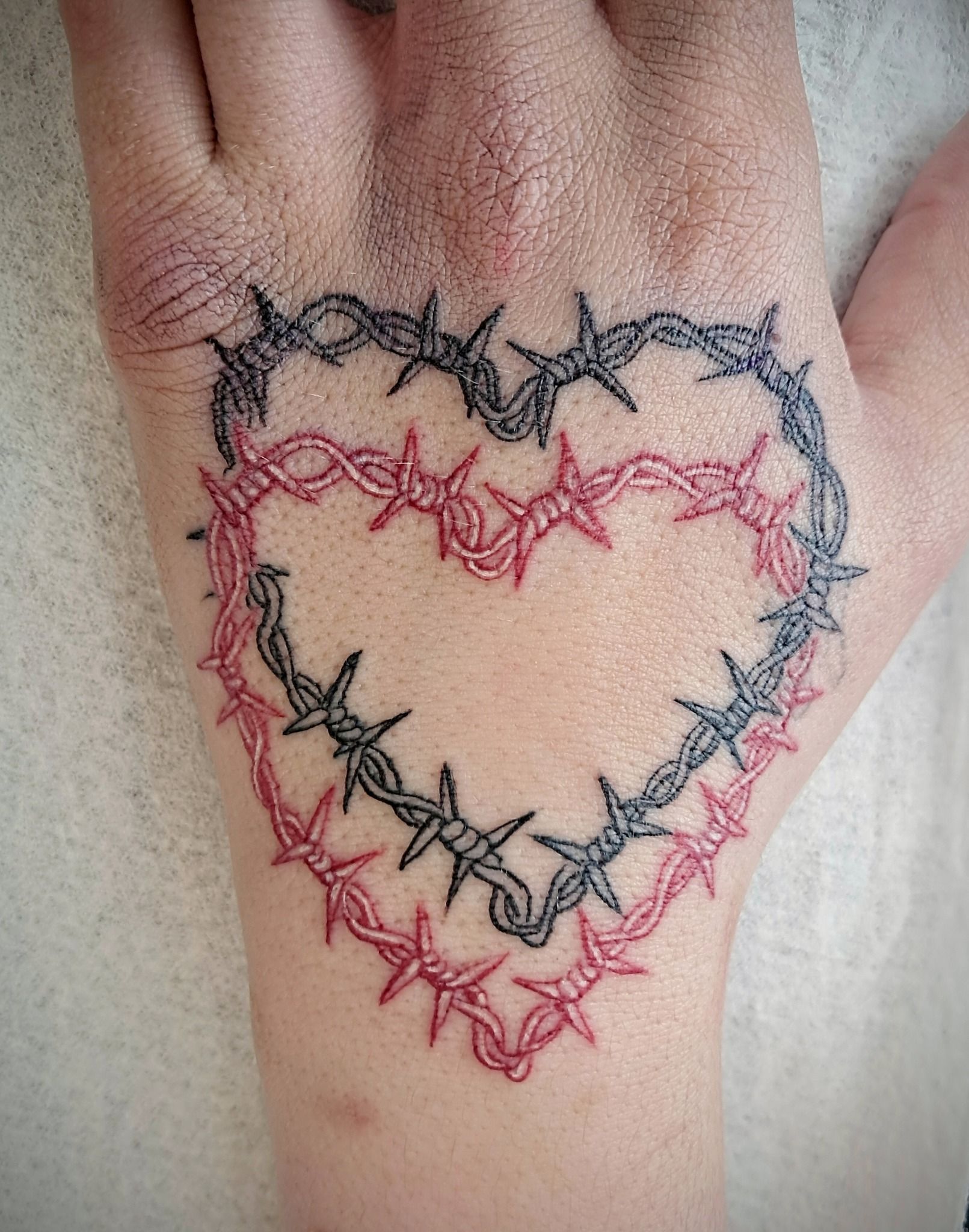 Tattoo of two barbed wire hearts on a hand. Red and black wire outlines the hearts.