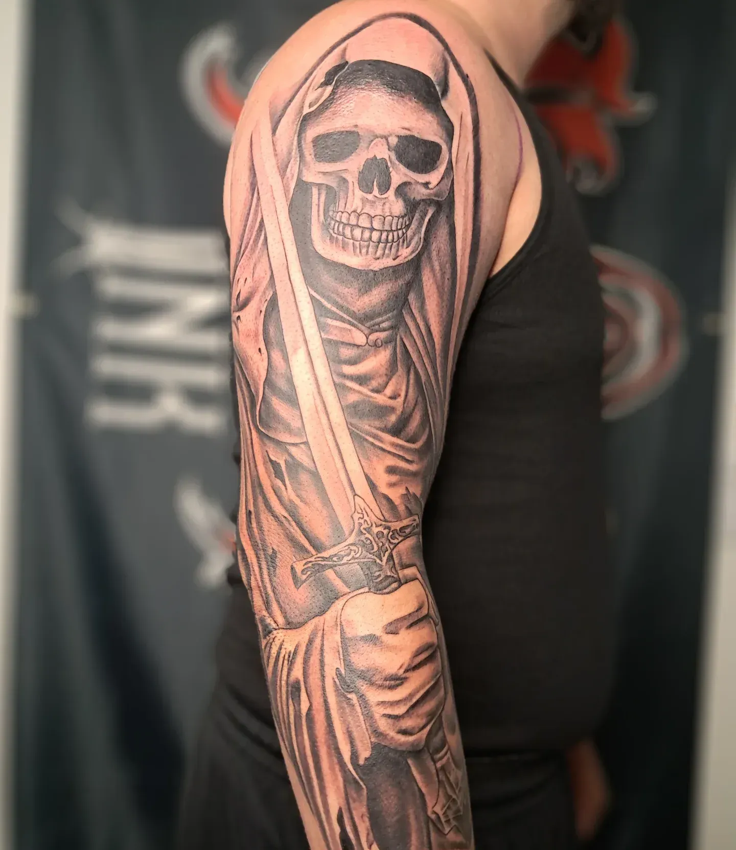 Tattoo of a skull wearing a hood holding a sword, on a person's arm. Black ink, grayscale shading.