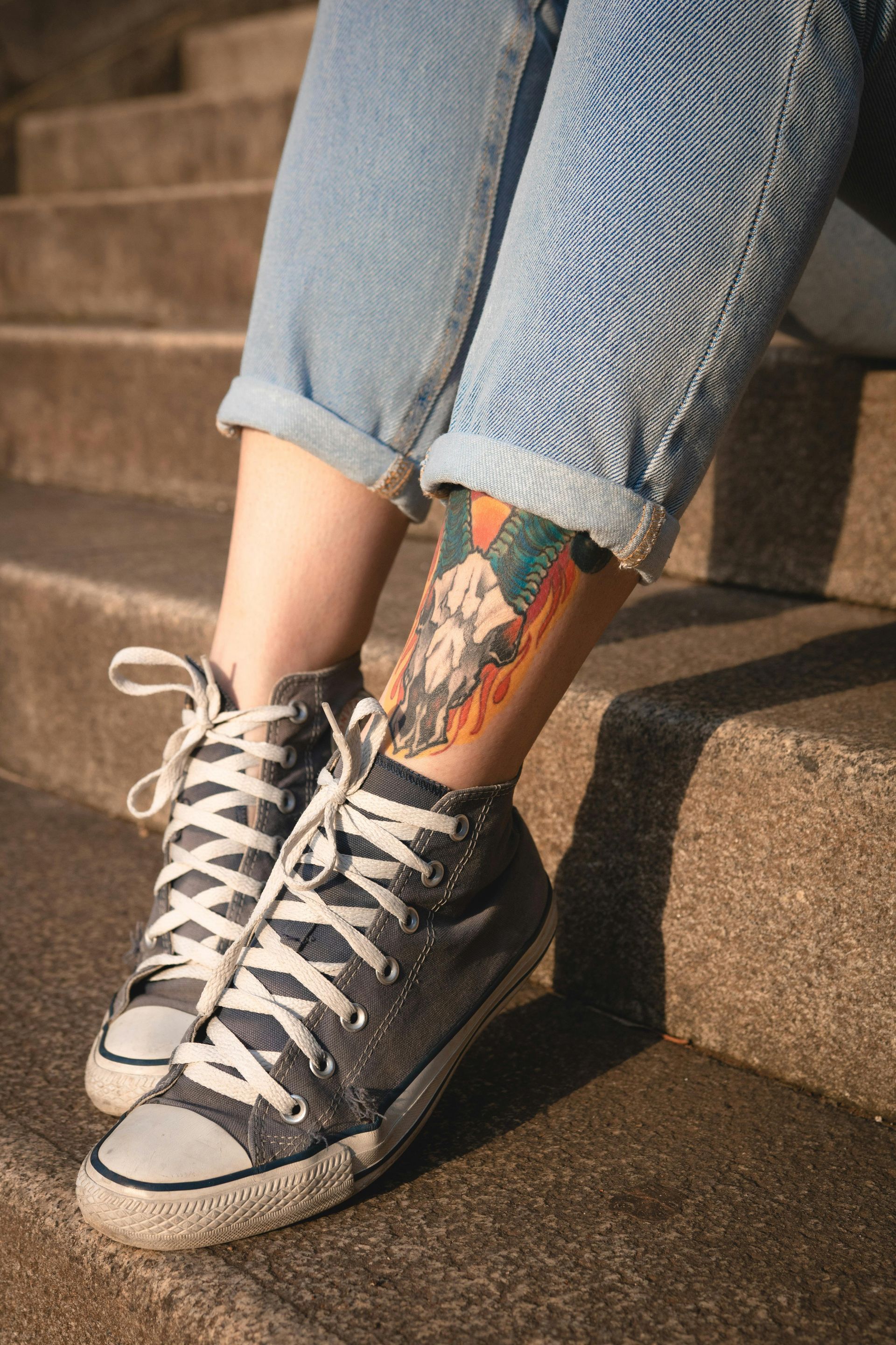 Person in jeans and high-top sneakers, sitting on steps with a leg tattoo visible.