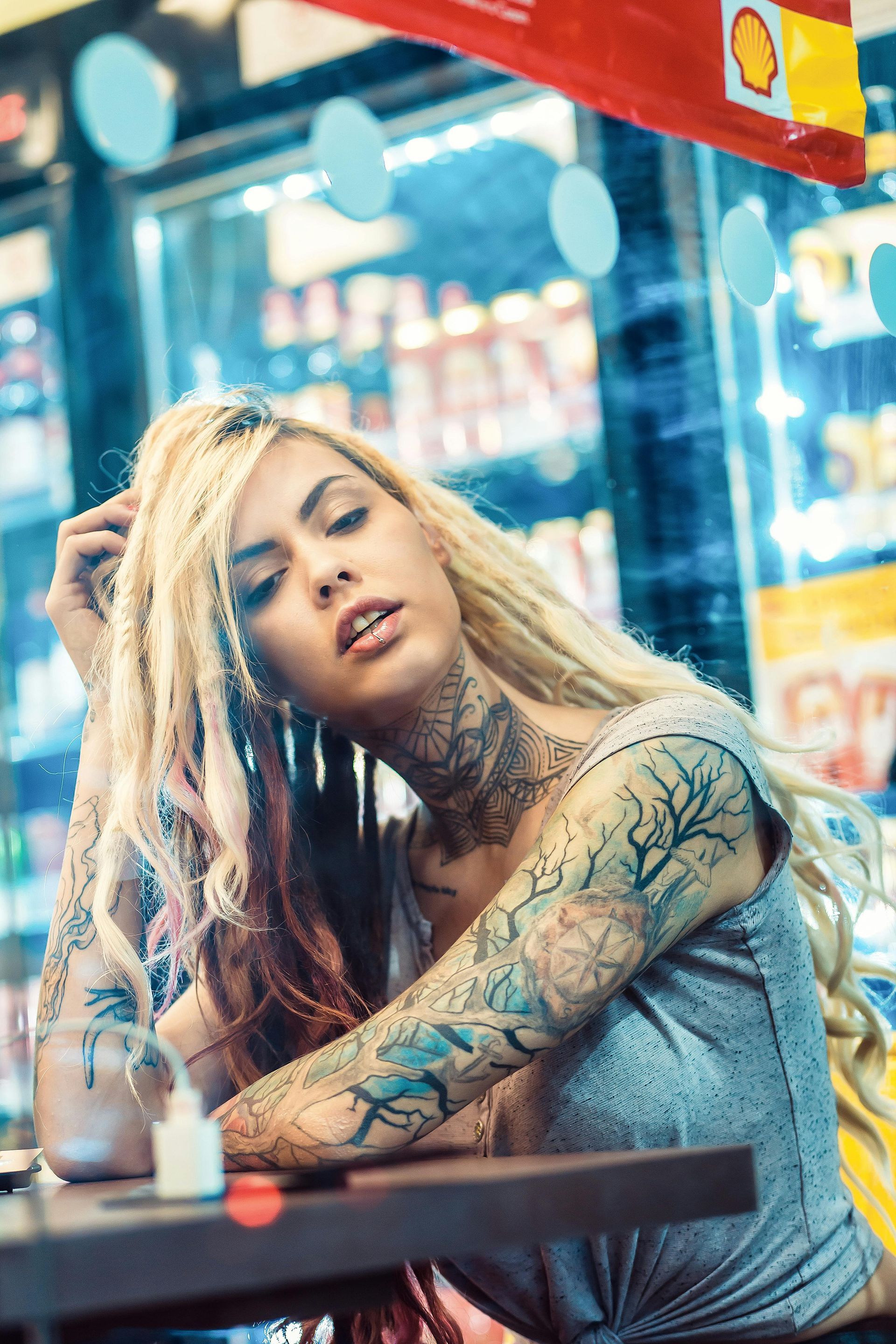 Blonde woman with tattoos at a store counter, looking up; arms on table.