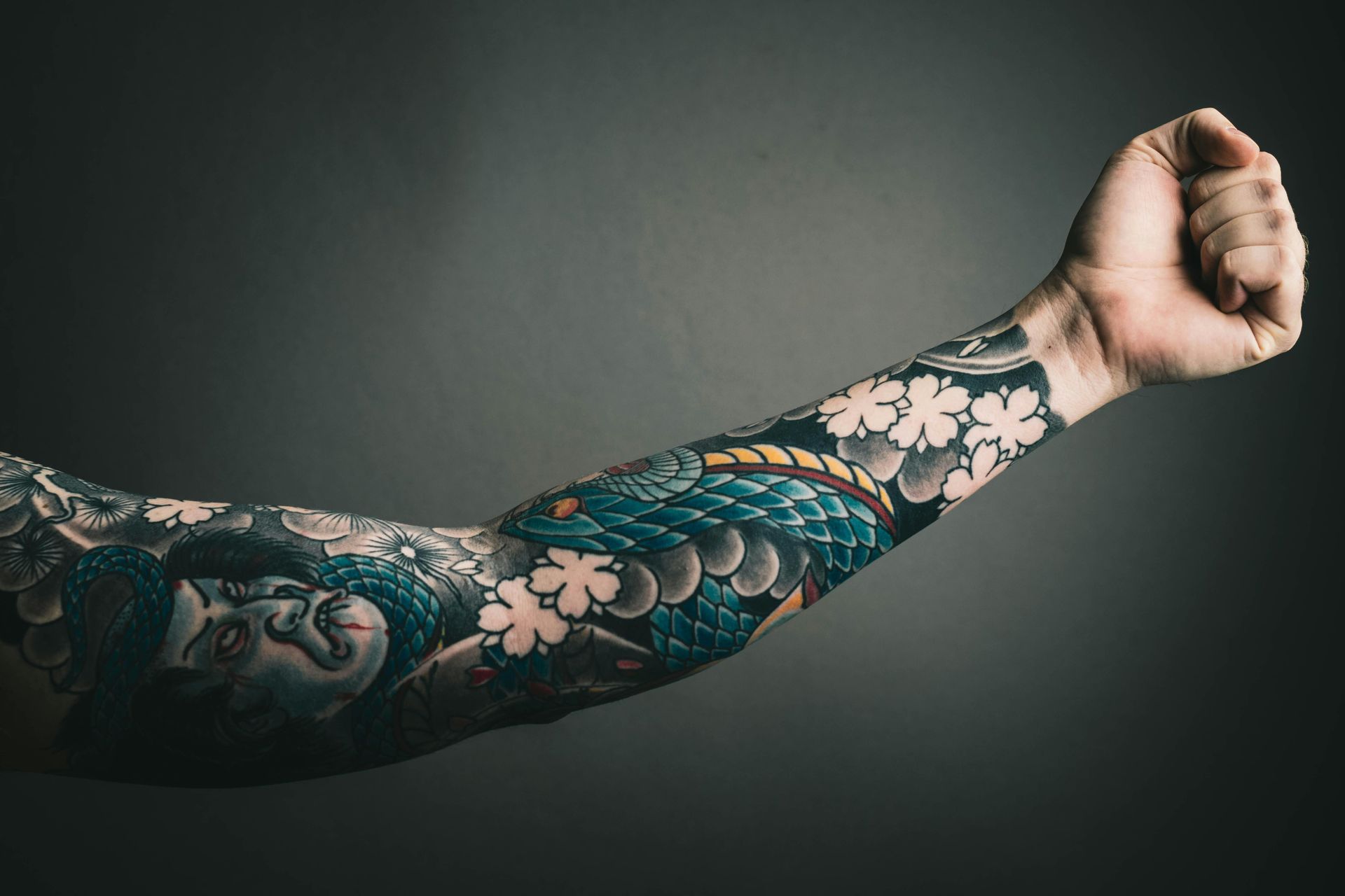 Arm covered in intricate blue, white, and black Japanese-style tattoos; clenched fist, gray background.
