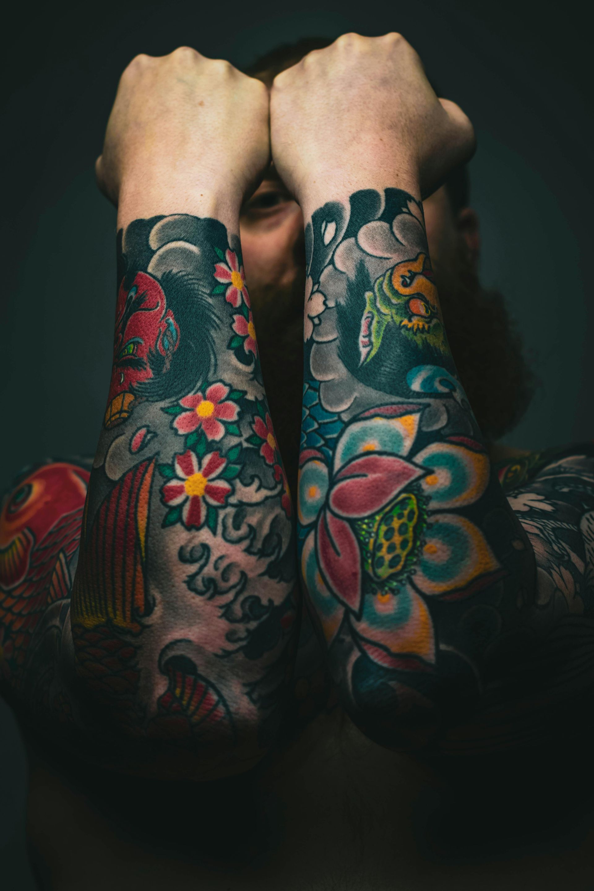Man with full sleeve tattoos, arms raised, hands near face. Colorful floral and wave designs.