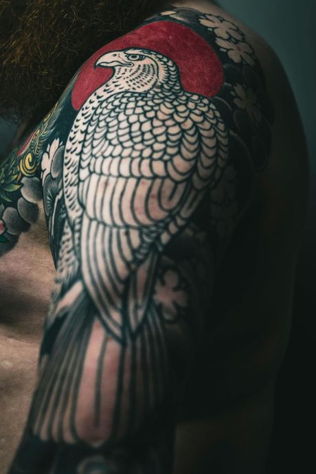 Tattooed shoulder with a detailed white and black eagle design, red sun, and floral elements.