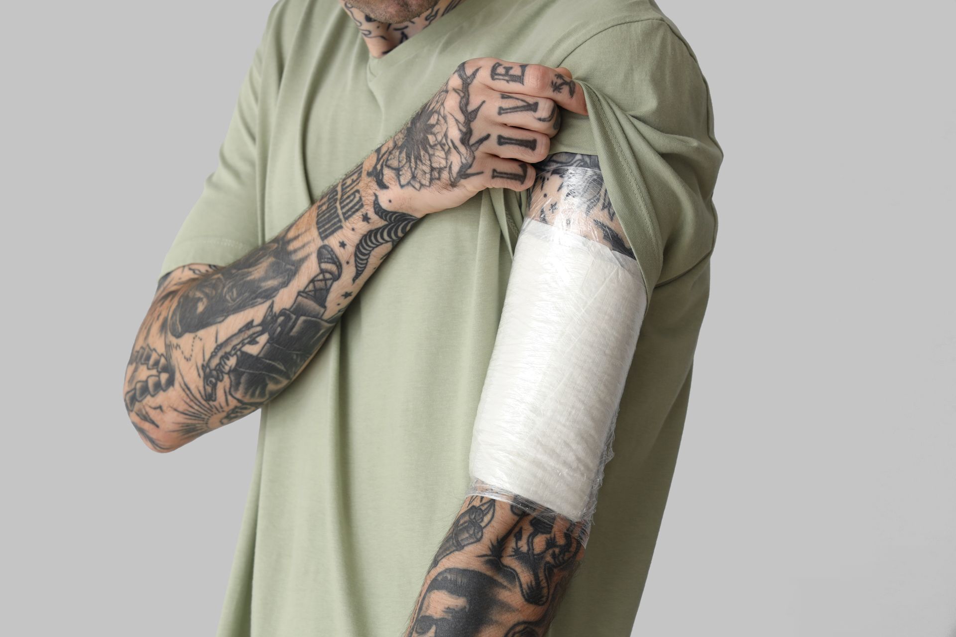 Person with tattoos, bandages on arm, pulling up sleeve against gray background.