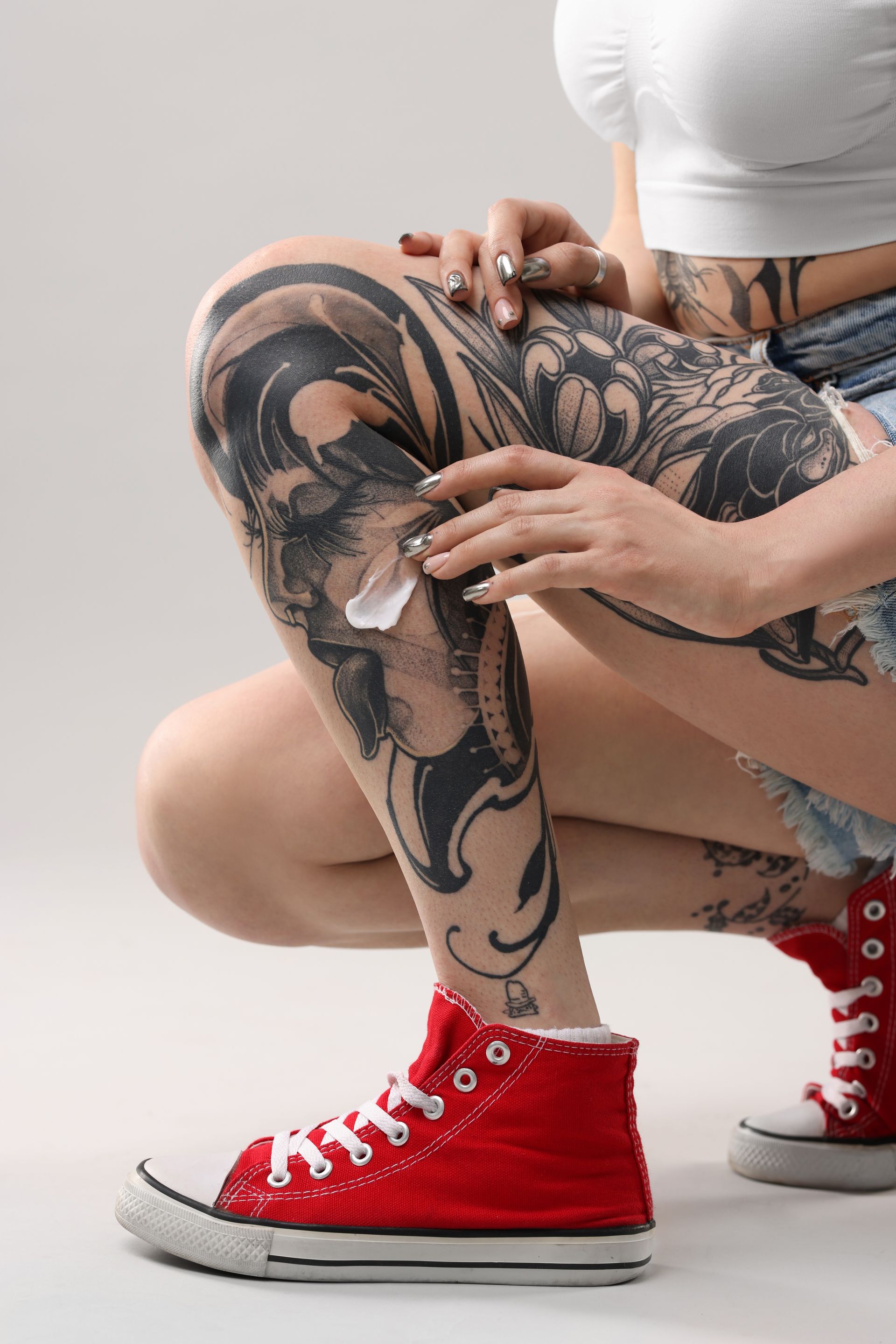 Woman with leg tattoos applying cream, wearing red sneakers.