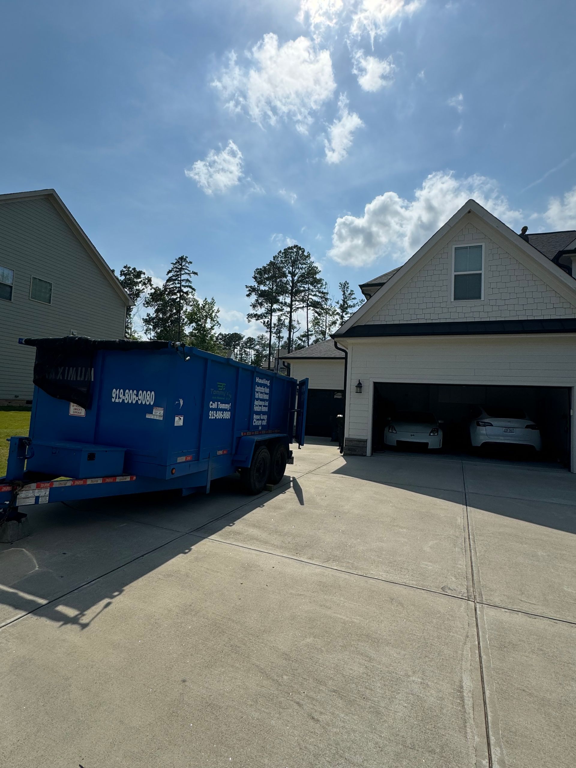 Blue generator trailer on a driveway, two cars in an open garage, blue sky with clouds.