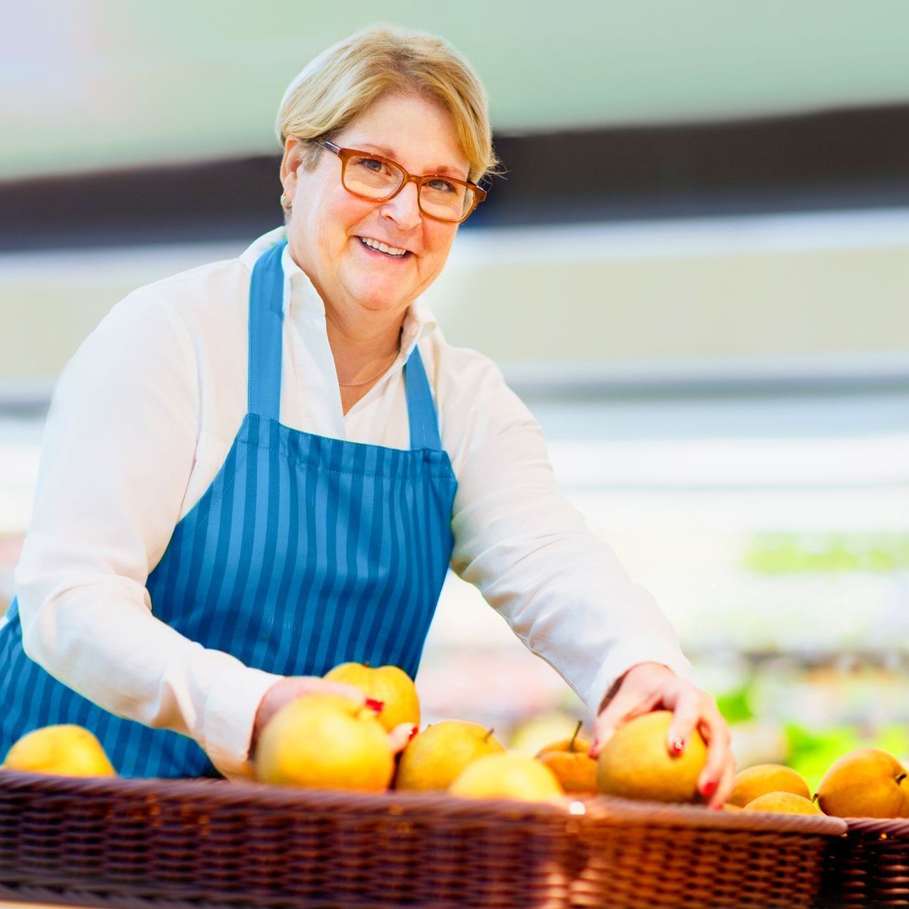 A woman in a blue apron is standing is re-stocking a basket of apples.
