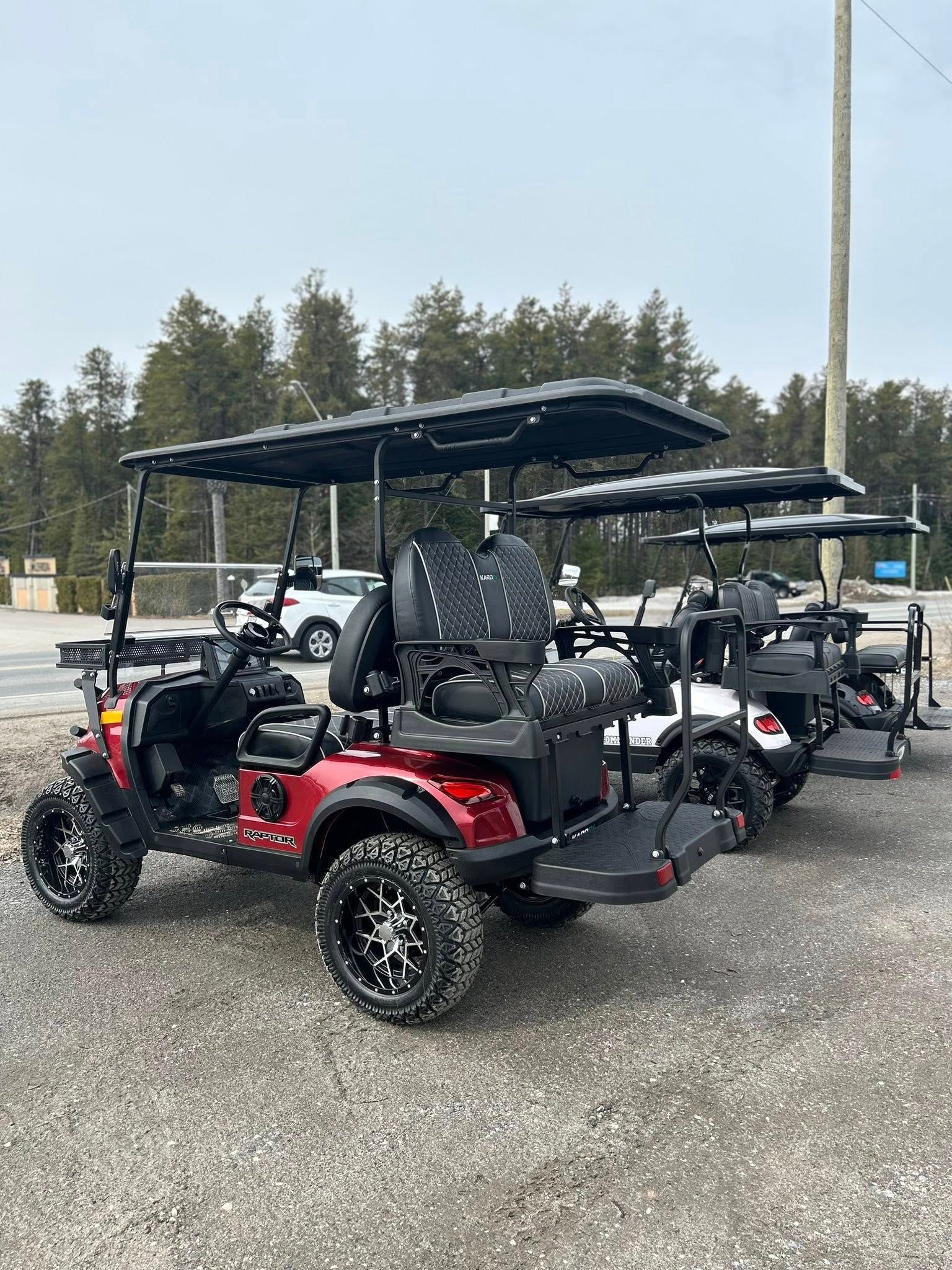 Voiturette de golf rouge avec toit noir et pneus tout-terrain garée sur du gravier avec deux autres, près des arbres et d'une route.