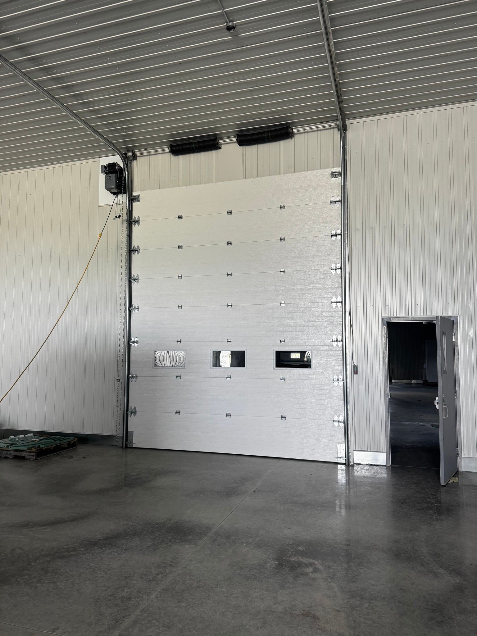 Vue intérieure d'un entrepôt avec une grande porte blanche et une porte plus petite ouverte sur une pièce sombre.