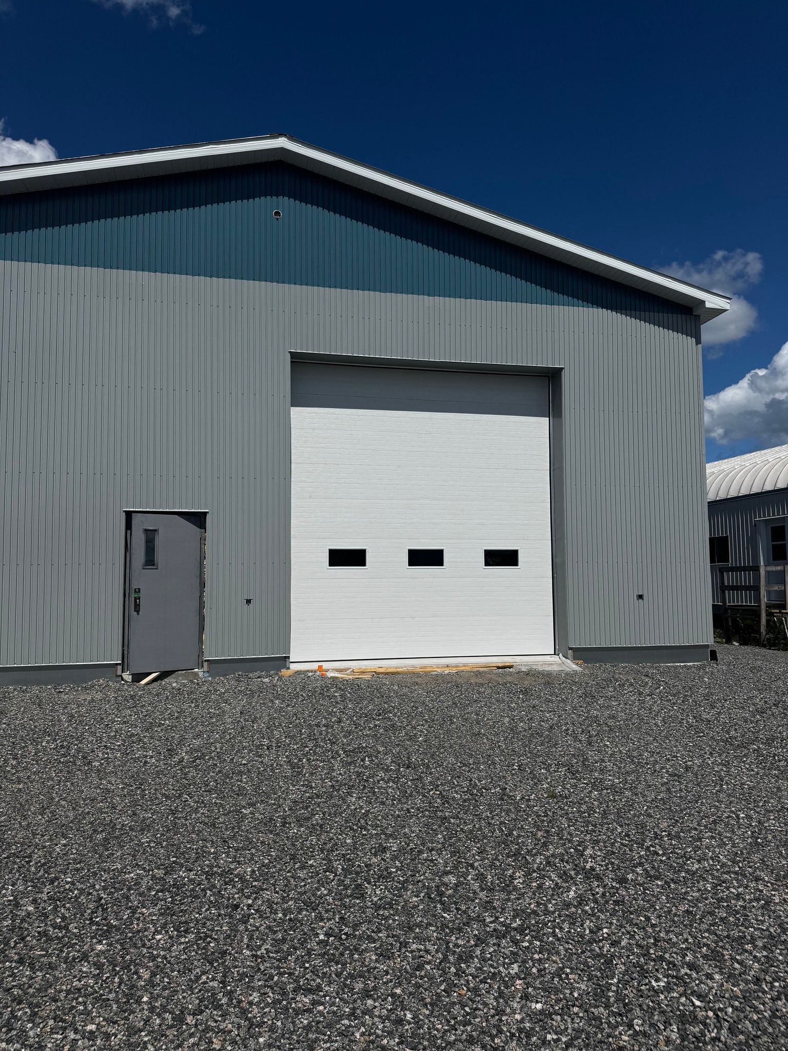 Bâtiment en acier avec porte de garage et porte latérale blanches sur gravier. Ciel bleu.