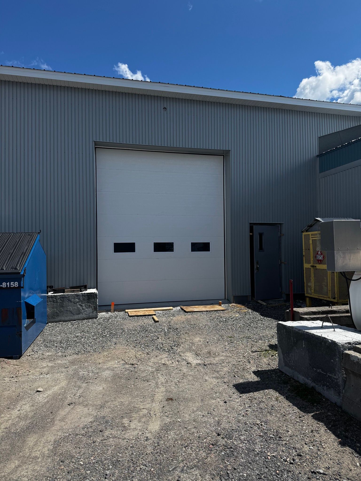 Vue extérieure d'une porte de garage blanche sur un bâtiment métallique. Une petite porte noire est adjacente.