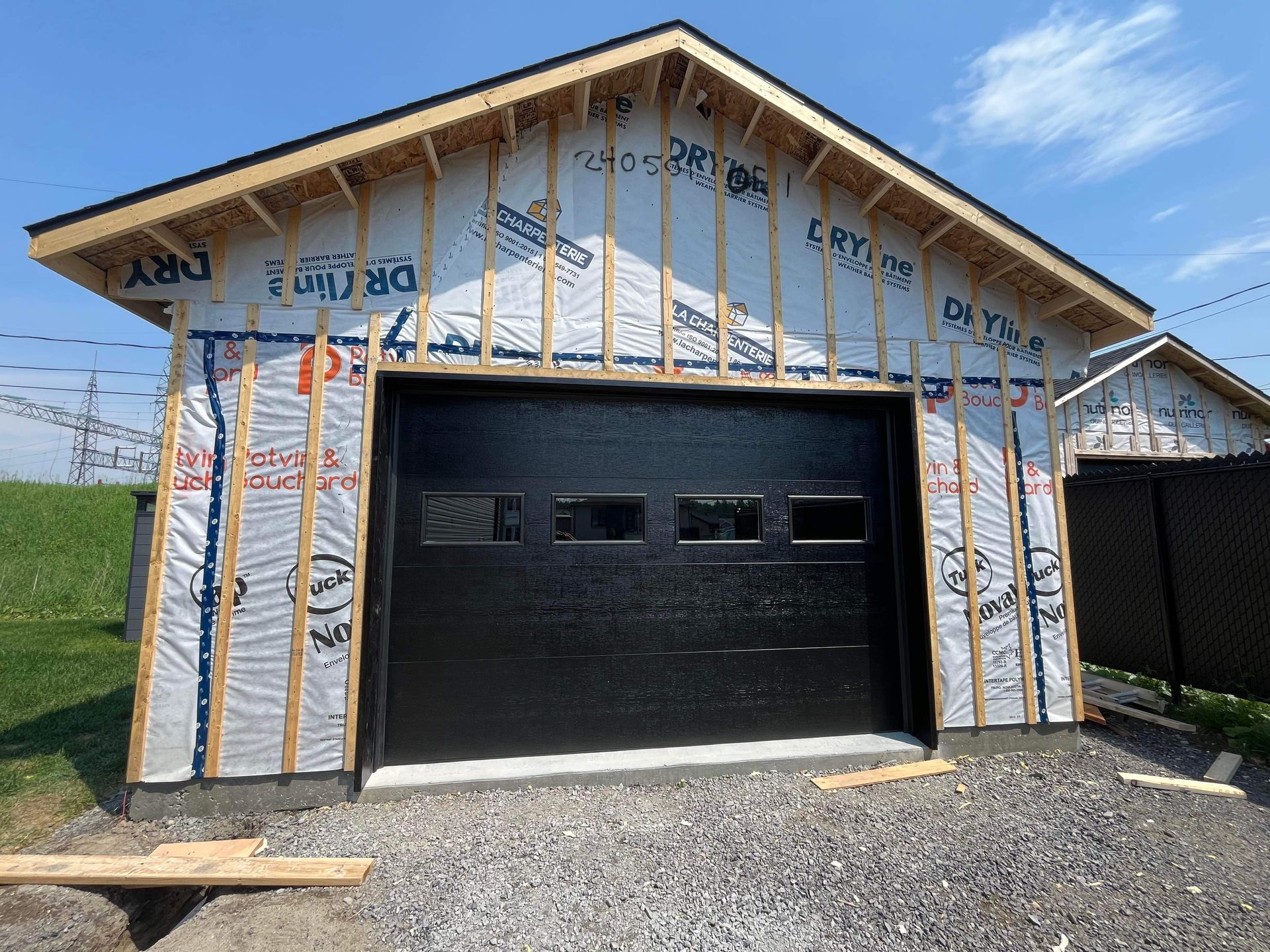 Porte de garage noire sur un immeuble en construction. Cadre en bois clair et revêtement bleu. Journée ensoleillée.