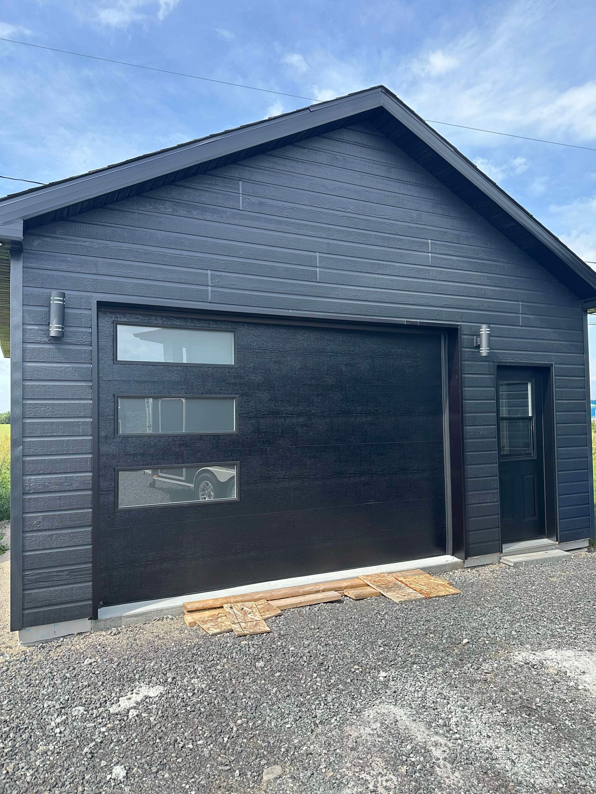 Garage noir avec une porte noire et des petites fenêtres; sol en gravier.