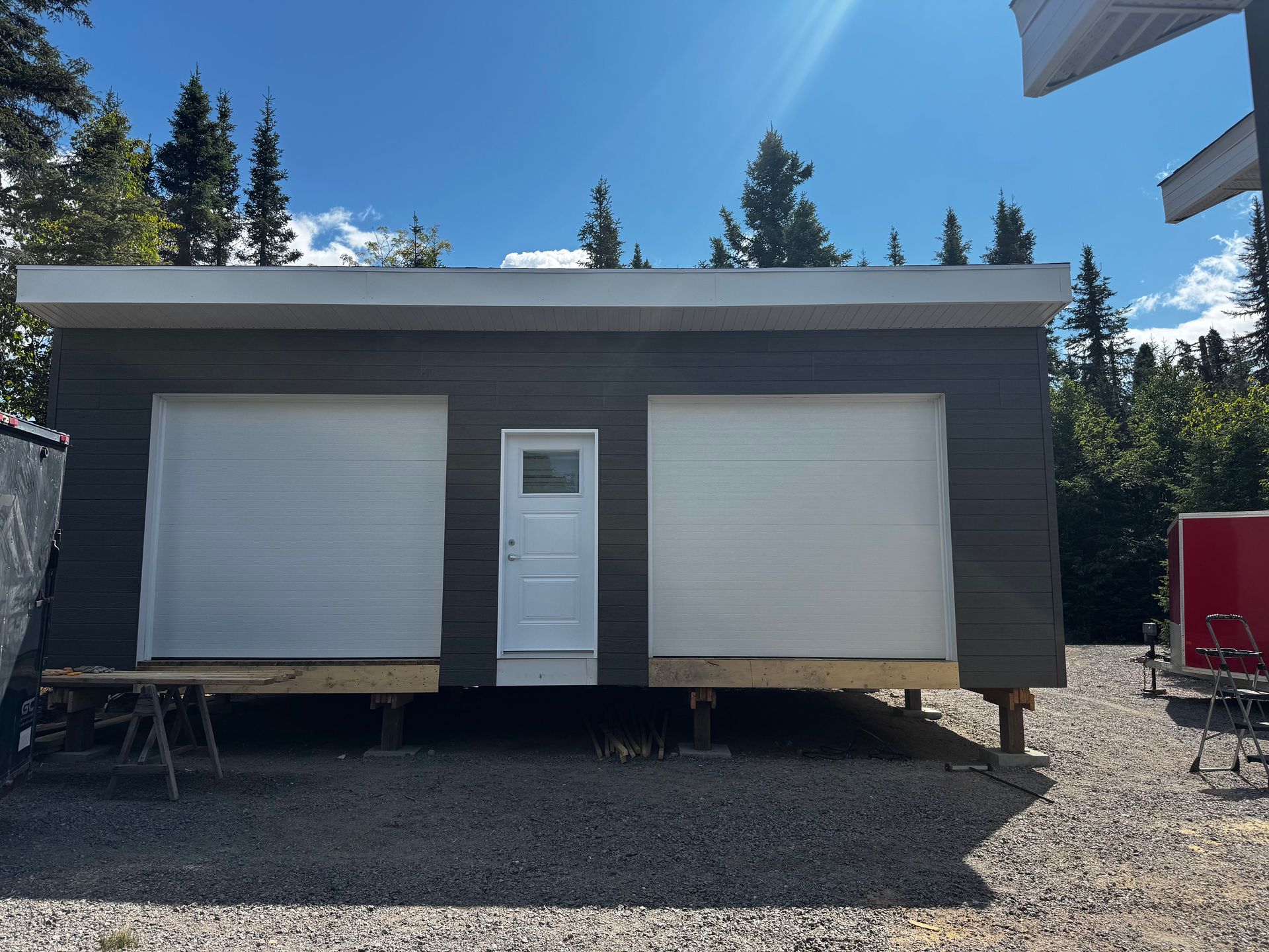 Bâtiment gris avec portes de garage blanches et une porte blanche centrée sur un terrain en gravier avec un fond de forêt.
