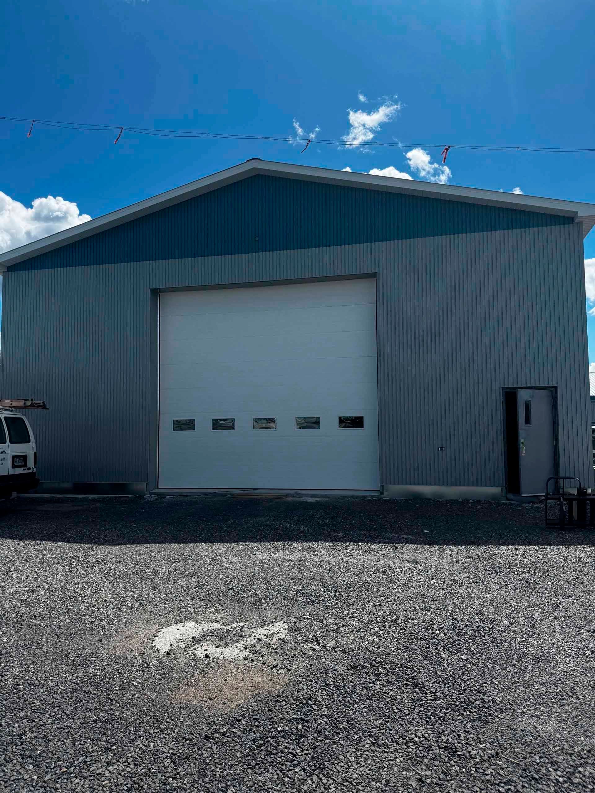 Bâtiment industriel métallique avec grande porte de garage, terrain gravillonné et ciel bleu.