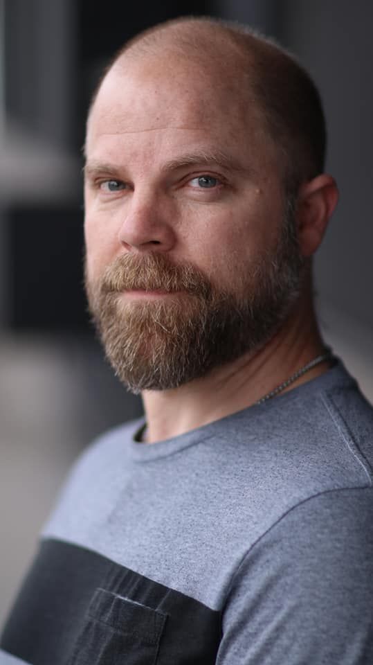 a bald man with a beard is wearing a striped shirt and looking at the camera .