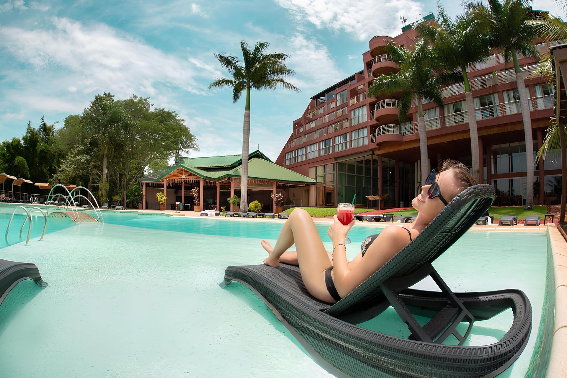 Una mujer está recostada en un sillón junto a una piscina sosteniendo una bebida.