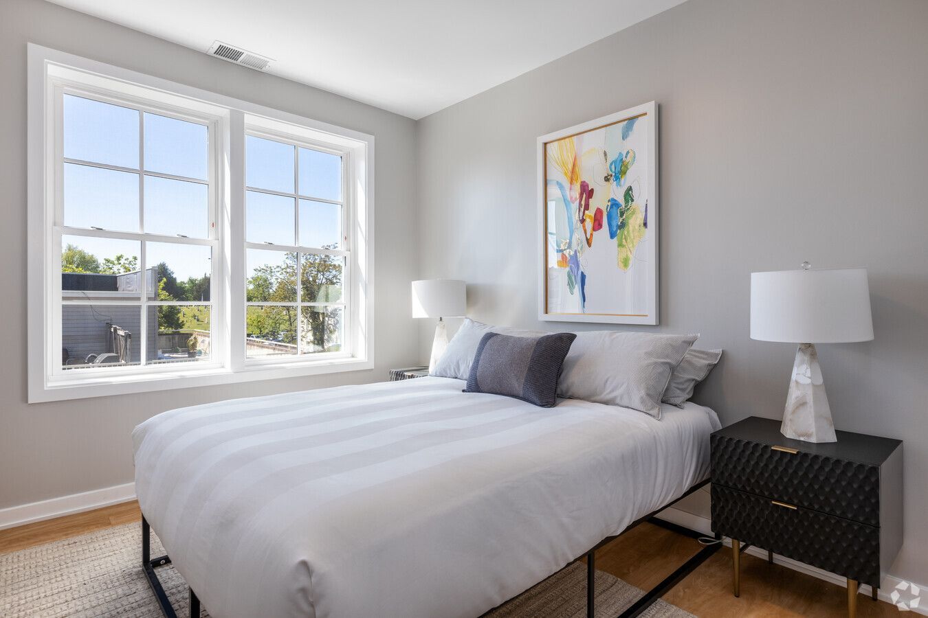 A bedroom with a bed , nightstand , lamps and two windows.