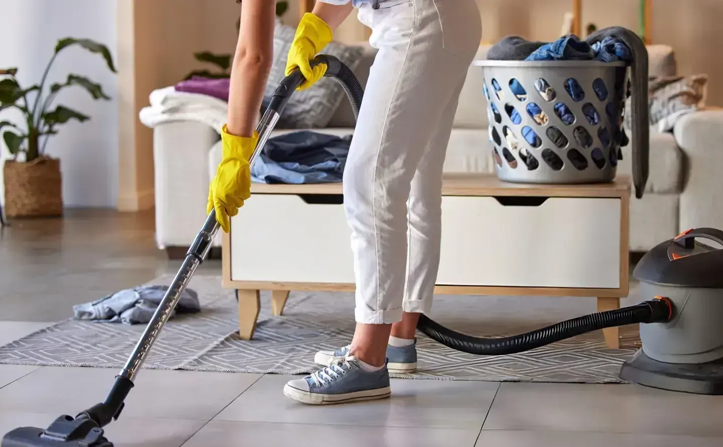 A professional cleaner in yellow gloves uses a steam extraction machine to clean a gray carpet in a living room.