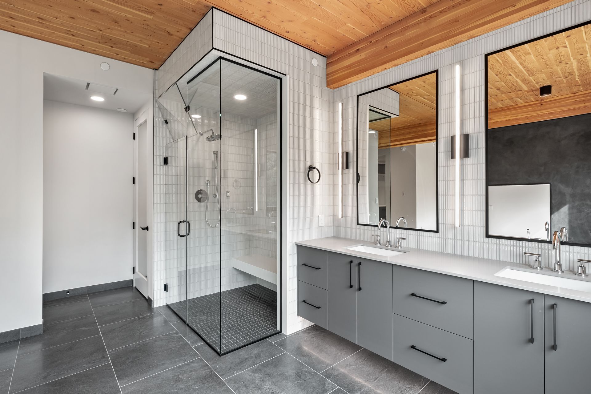 Modern bathroom with glass shower, gray vanity, wood ceiling, and dark tile floor.