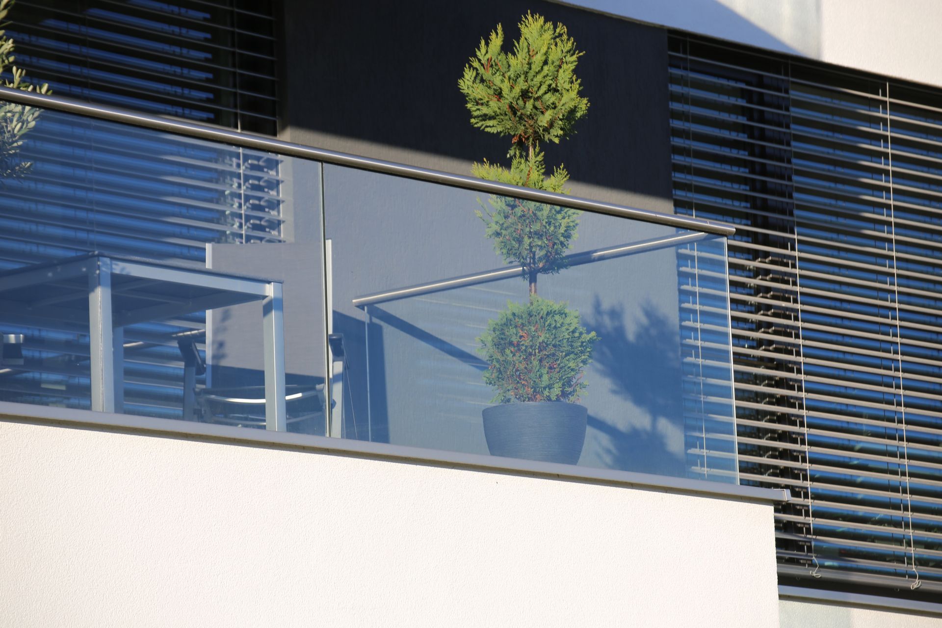 Modern balcony with glass railing, table, potted plants, and slatted sun shades.
