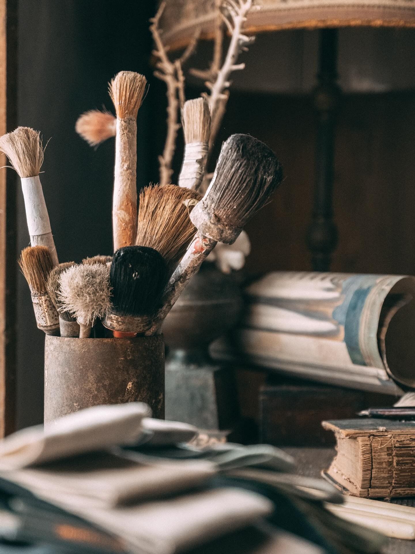 Pennelli in un contenitore di metallo arrugginito su uno scaffale ingombro di vecchi libri e carta arrotolata.