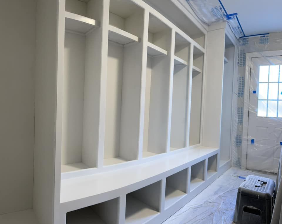 White built-in storage unit with cubbies, shelves, and bench seat in a room. Door and work tools are in background.