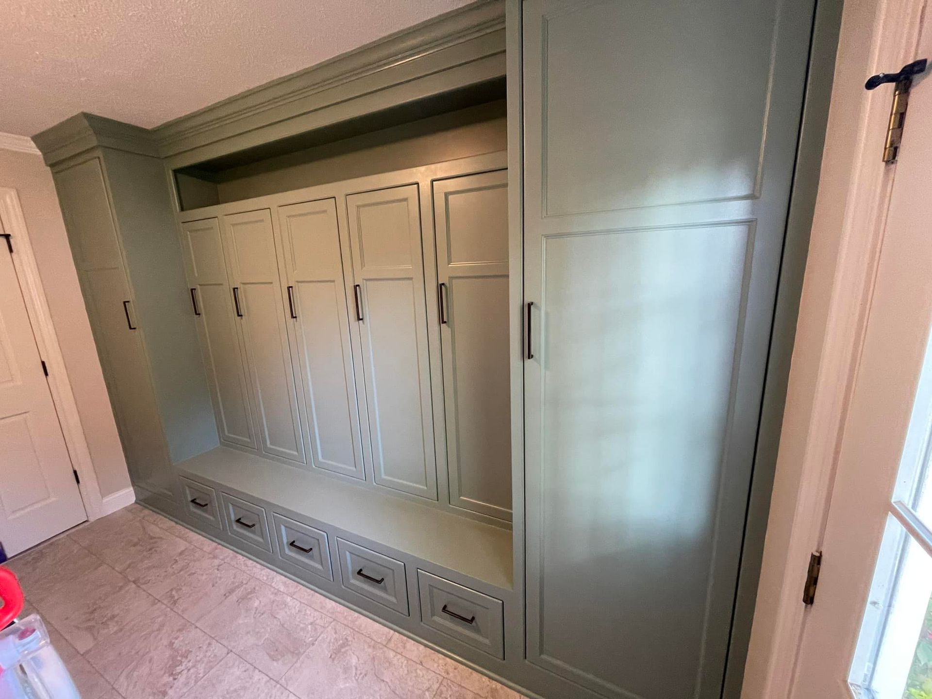 Built-in green cabinet with hooks, doors, drawers, and a bench, in a room with a window and door.