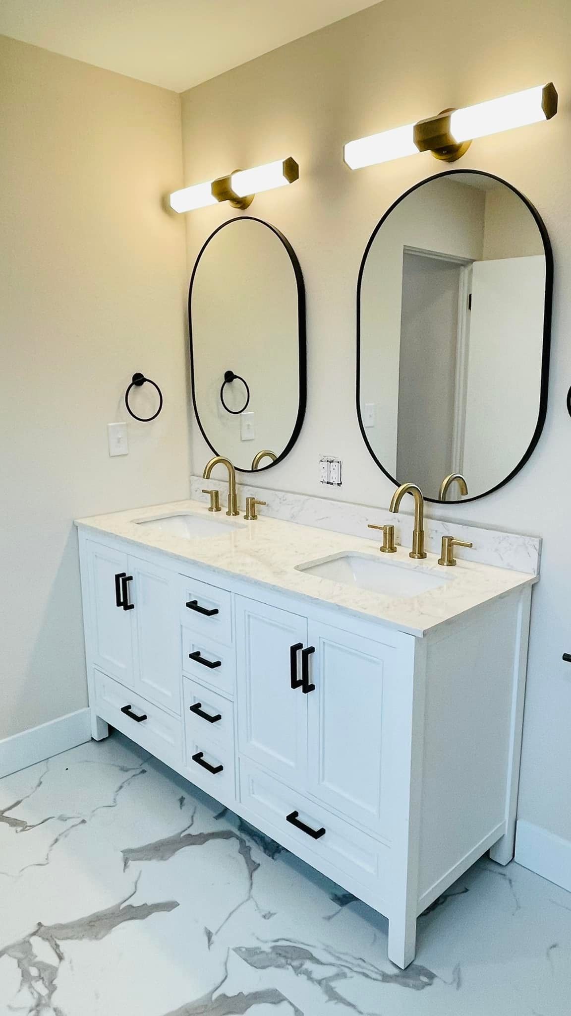 White double sink vanity with gold fixtures, oval mirrors, and marble-look tile.