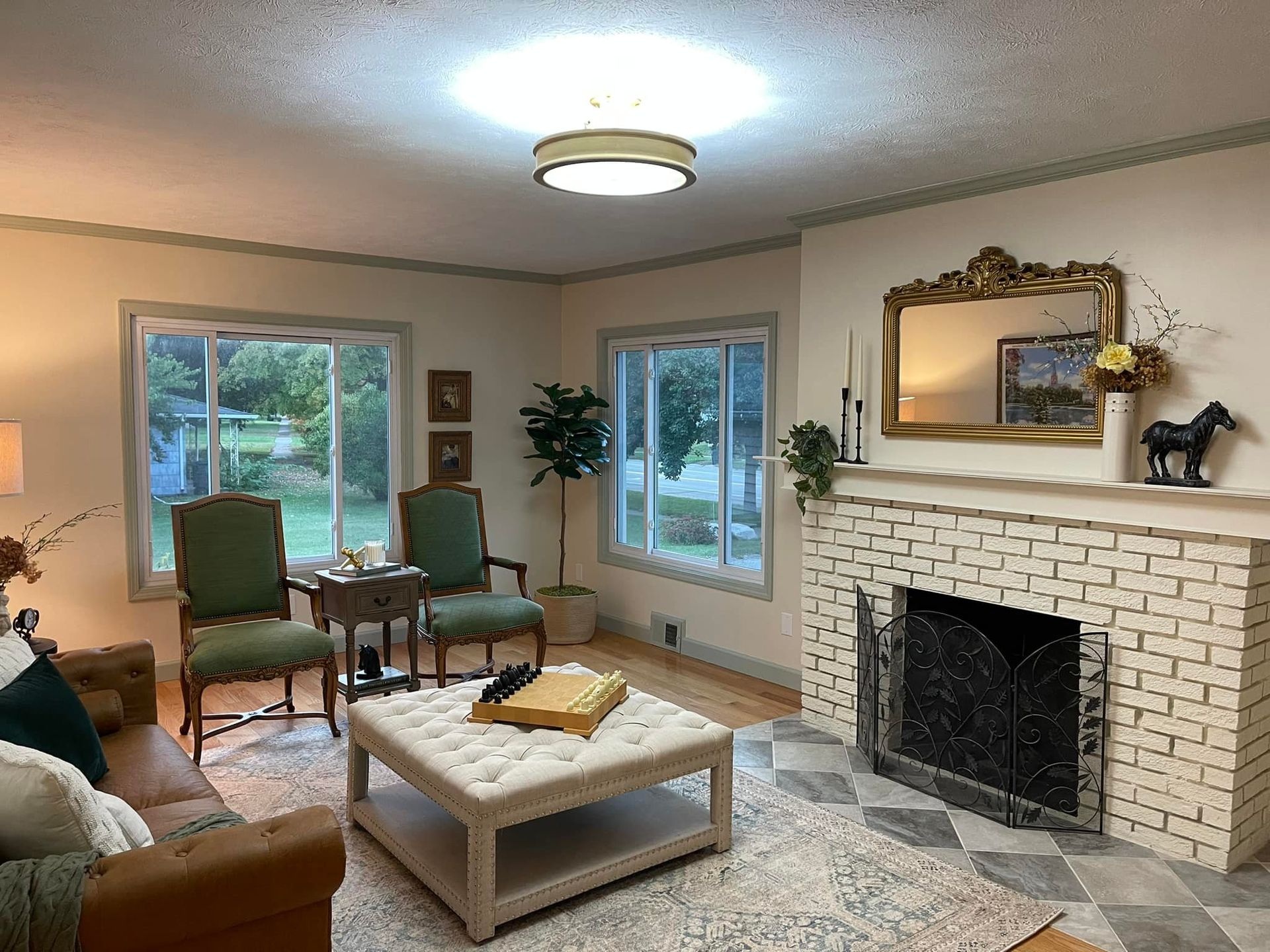 Living room with fireplace, two chairs by window, ottoman coffee table, and large ornate mirror.
