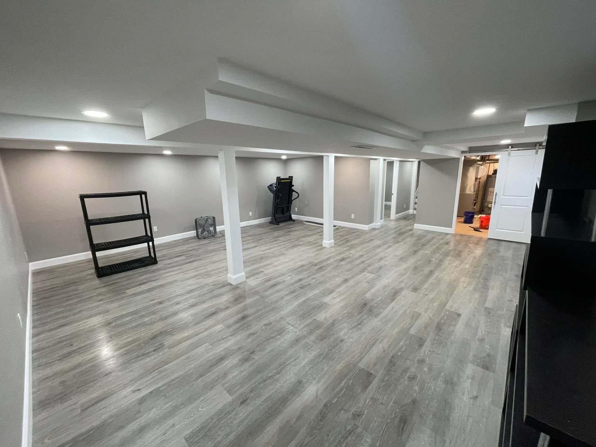 Empty, open basement with gray walls and wood-look flooring. Black shelving and gym equipment are present.