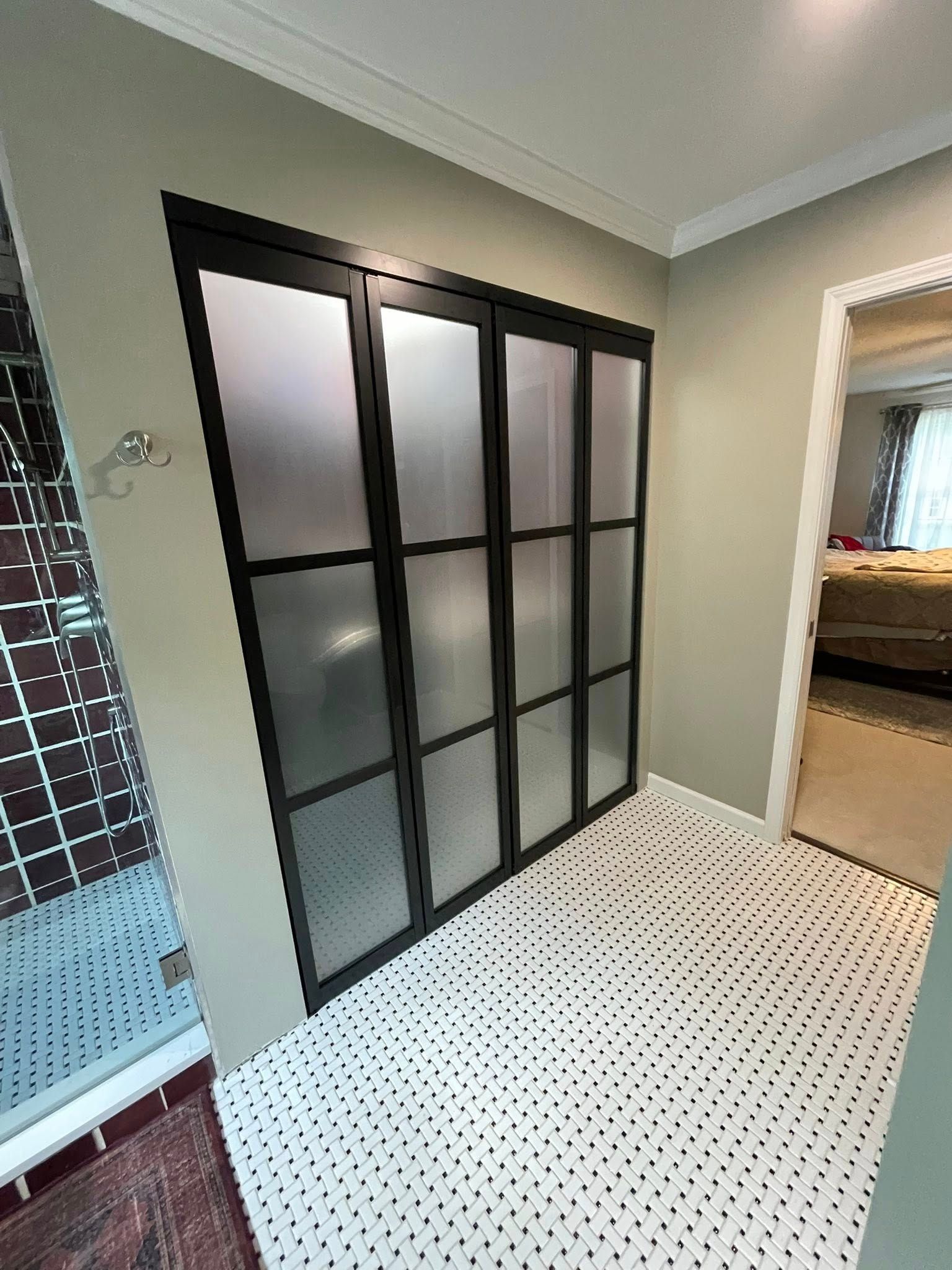 Bathroom with black-framed, frosted glass closet doors and white penny tile floor. Shower visible on the left.
